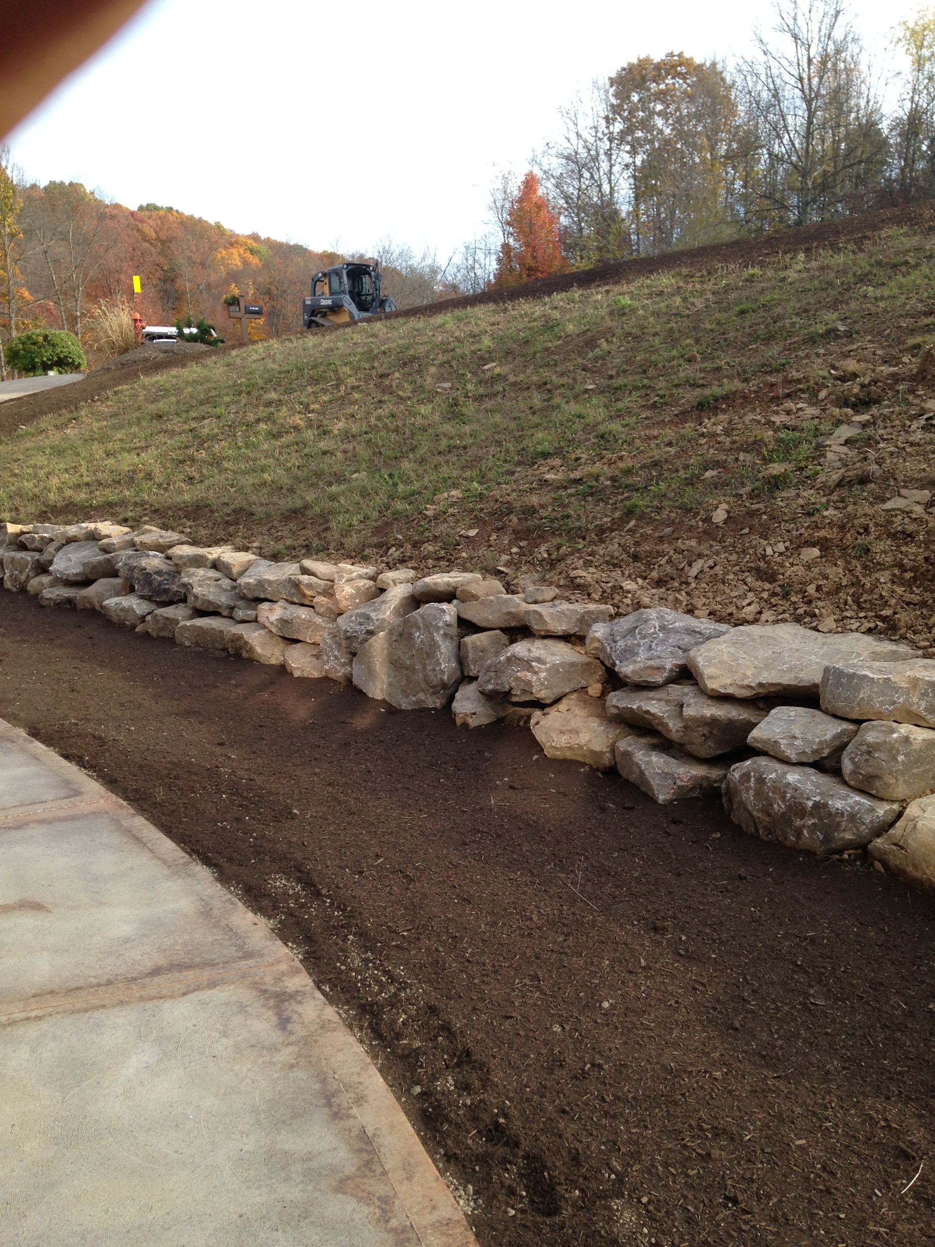 Curved stone retaining wall beside a sloped dirt embankment and paved path, with autumn trees in the background