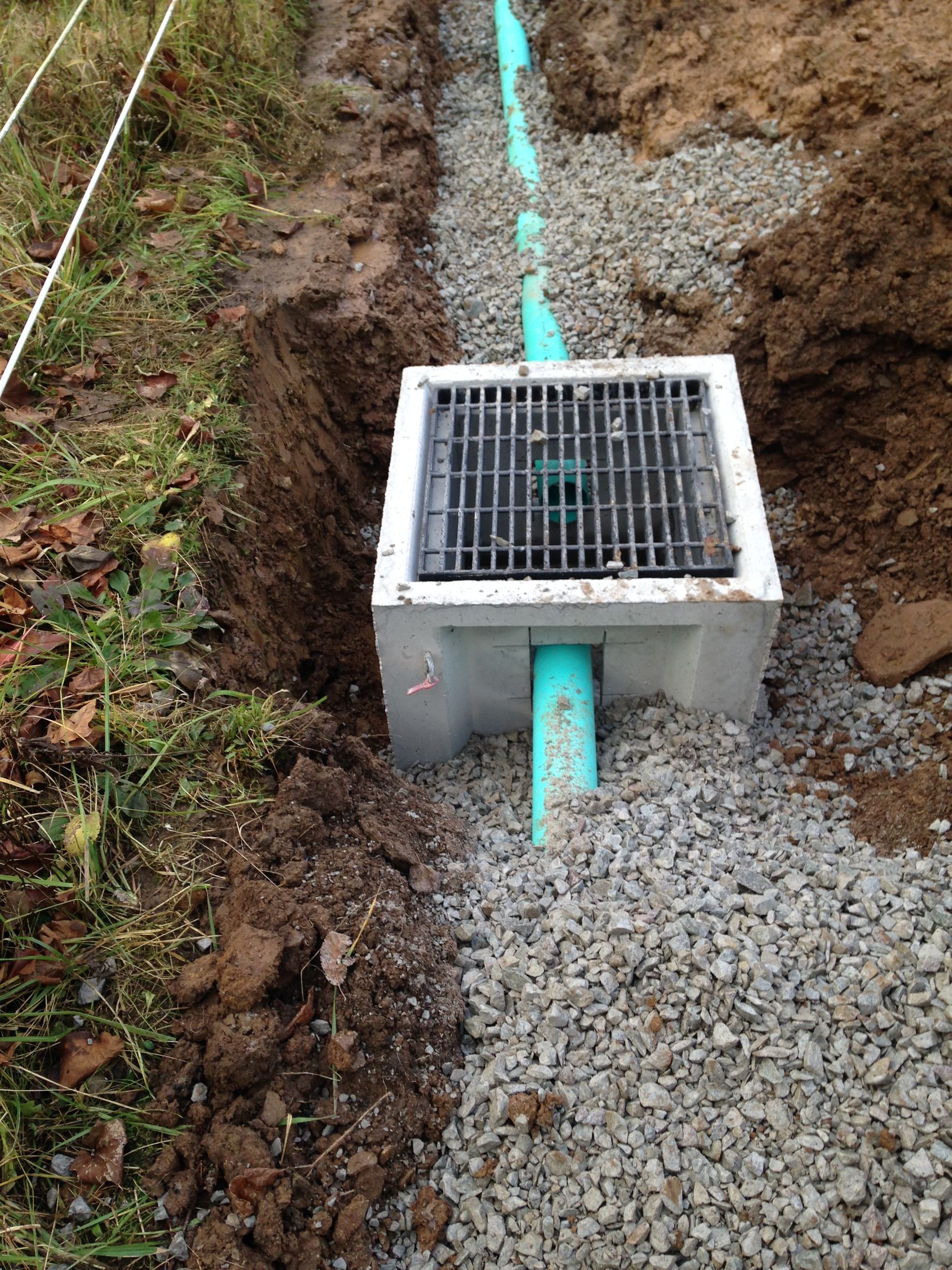 Small utility drain box with grate over a turquoise pipe in a gravel trench by a grassy edge