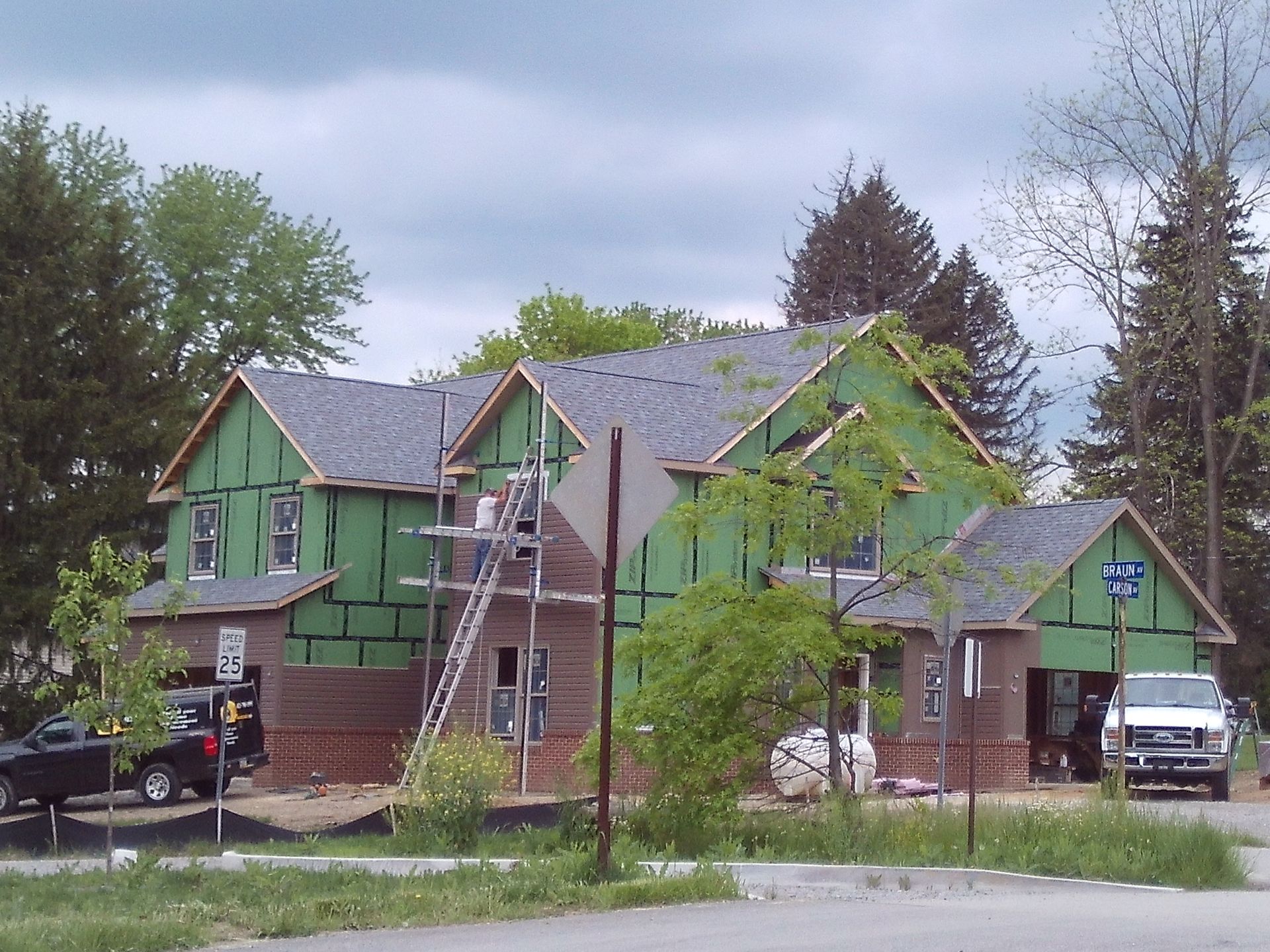 Green houses under construction in a wooded neighborhood, with scaffolding and trucks on the street