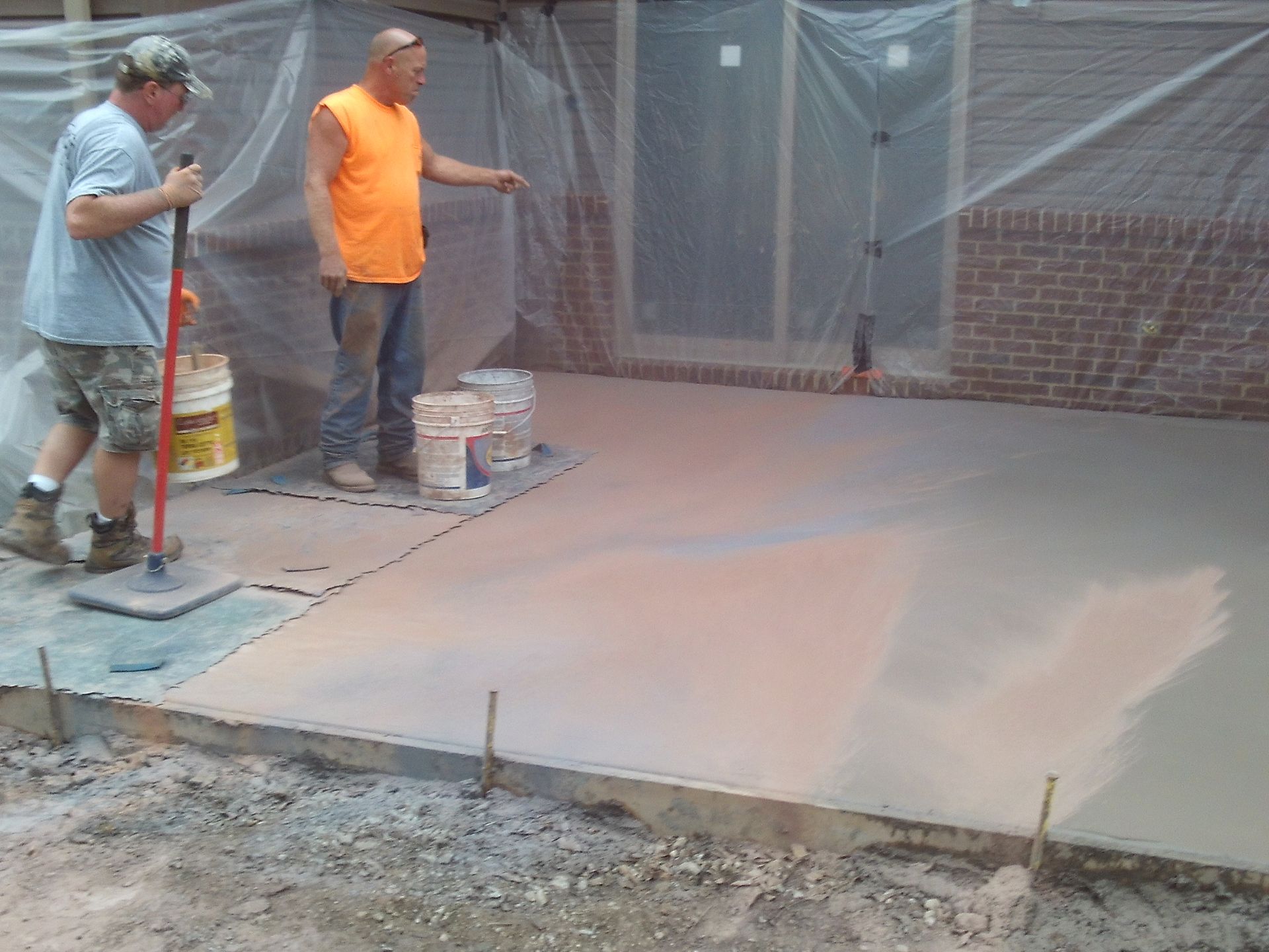 Two workers smoothing fresh concrete on a patio beside a brick wall and covered fencing