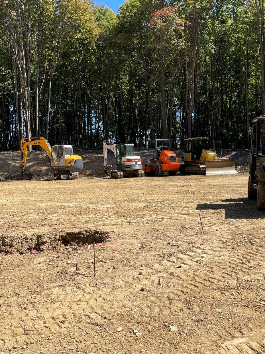 Construction equipment in a cleared wooded area, including an excavator, skid steer, and orange utility vehicle.