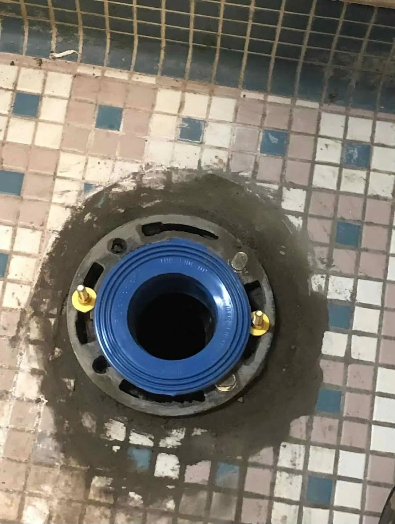 Blue toilet flange on tiled bathroom floor with exposed bolts and rough concrete around it