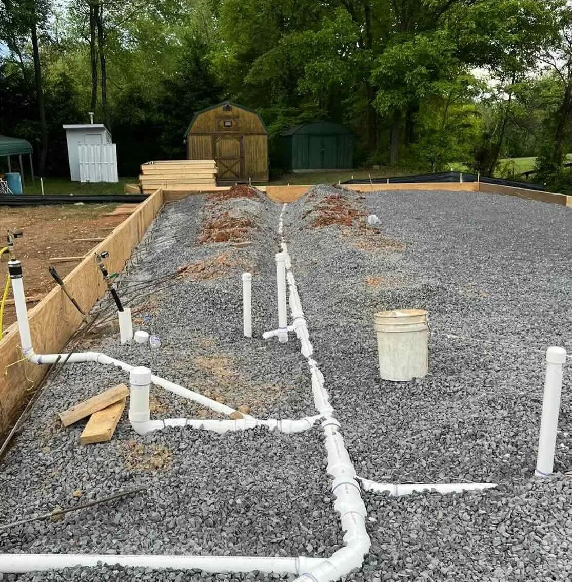 Gravel construction site with white PVC pipes and trenches, trees and a shed in the background