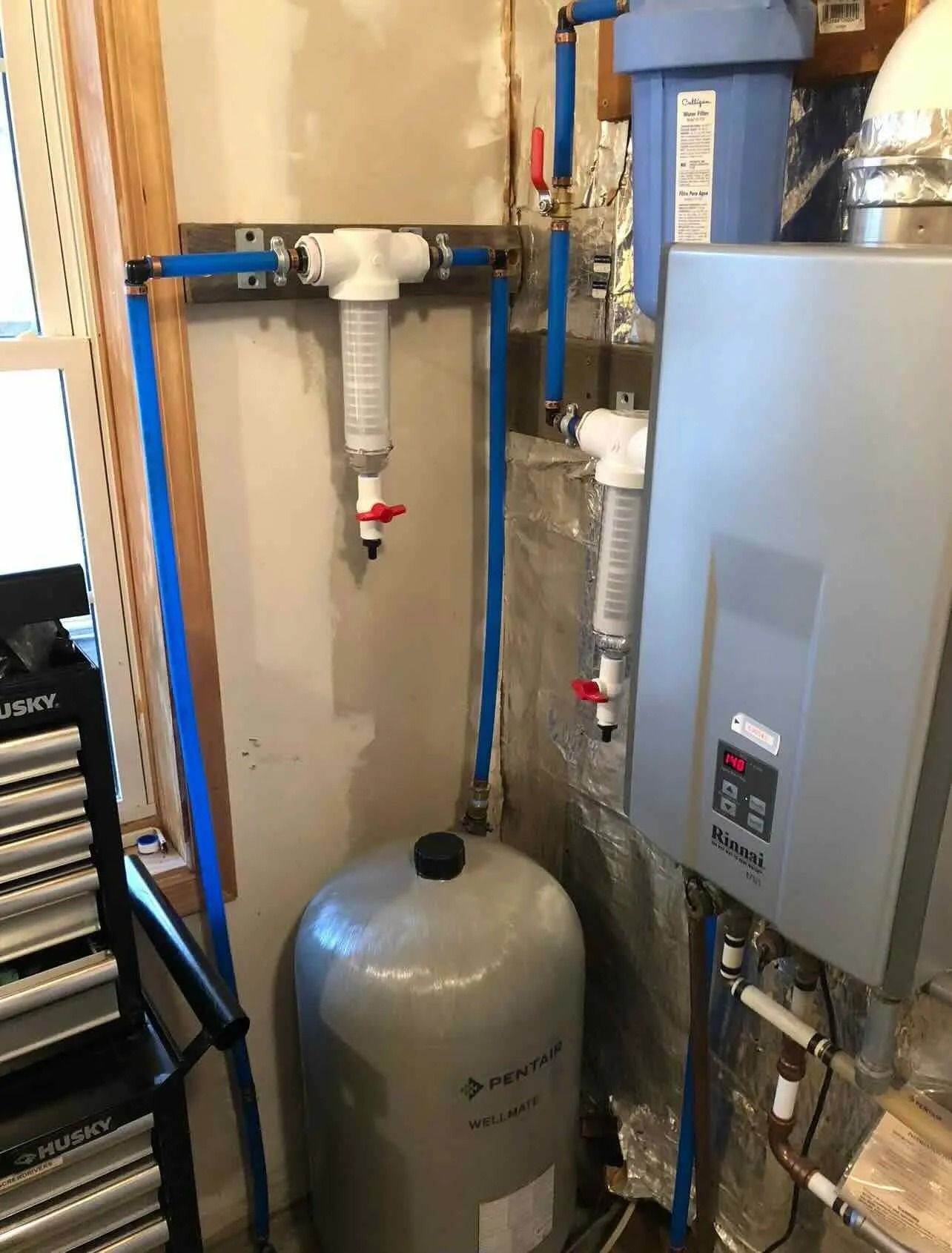 Basement utility area with plumbing pipes, water tank, and gray boiler unit.