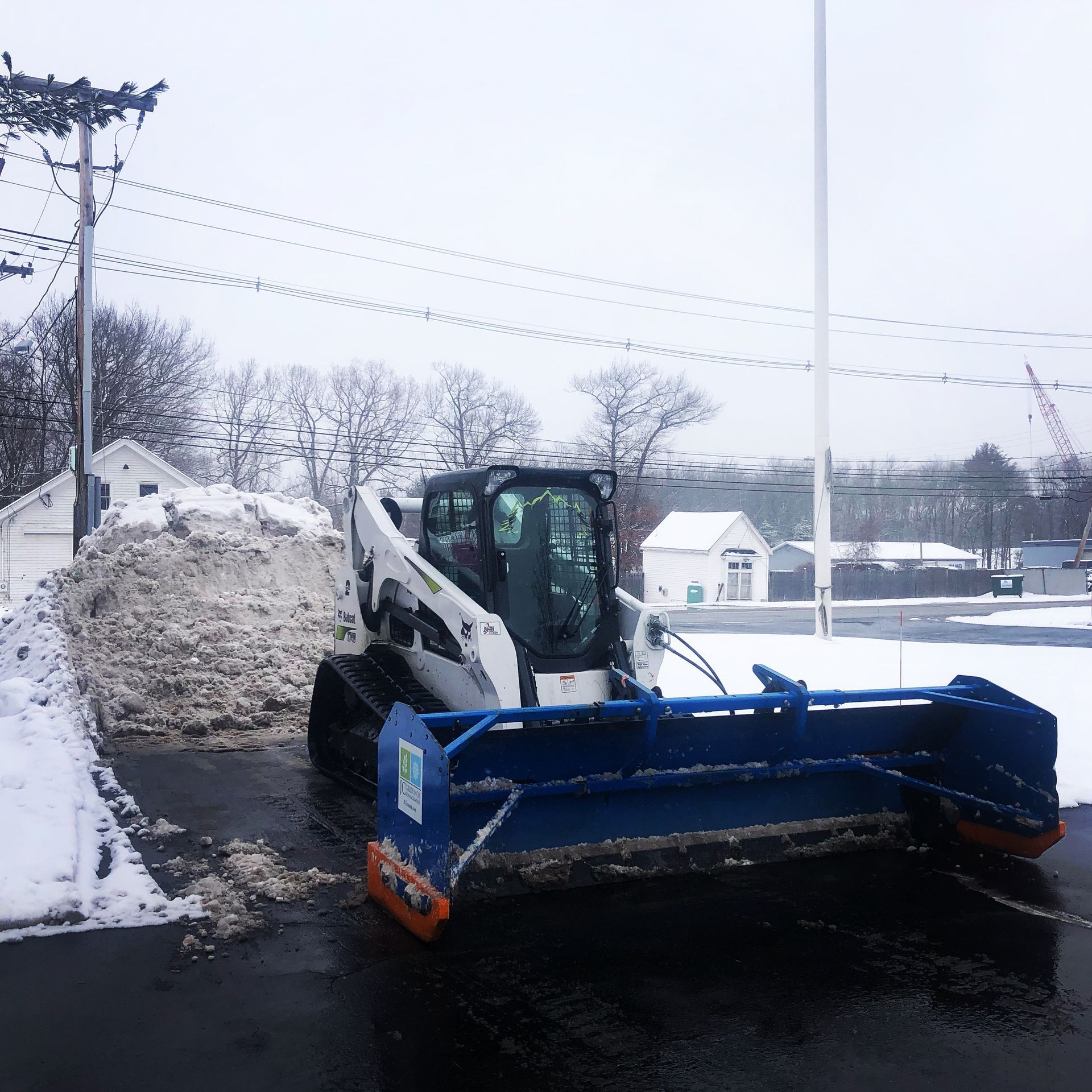 A white and black skid-steer loader with a large blue snow plow attached, parked on wet pavement next to a snow pile.