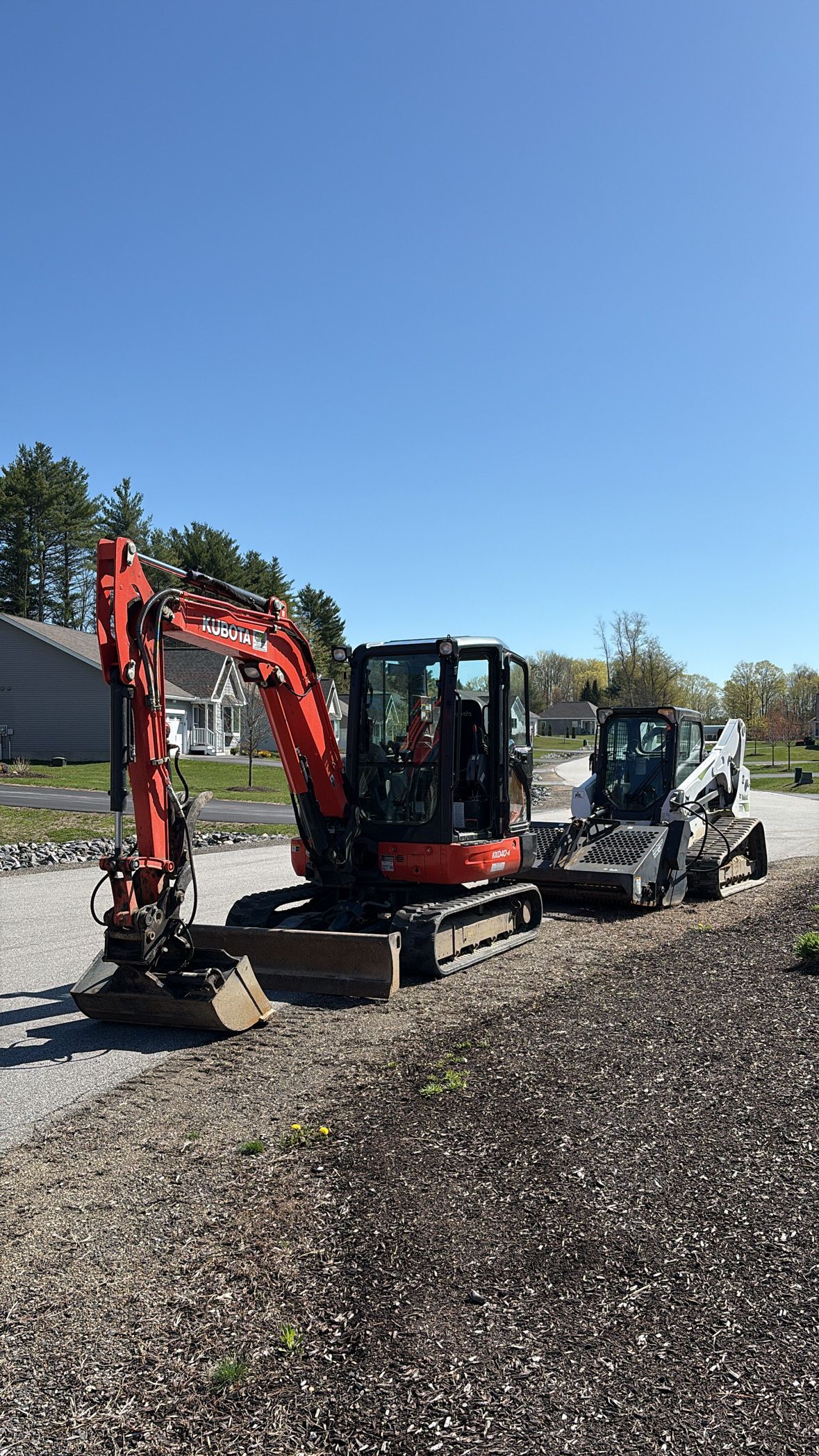 An orange mini-excavator and a white skid steer loader parked on a gravel path under a clear blue sky.