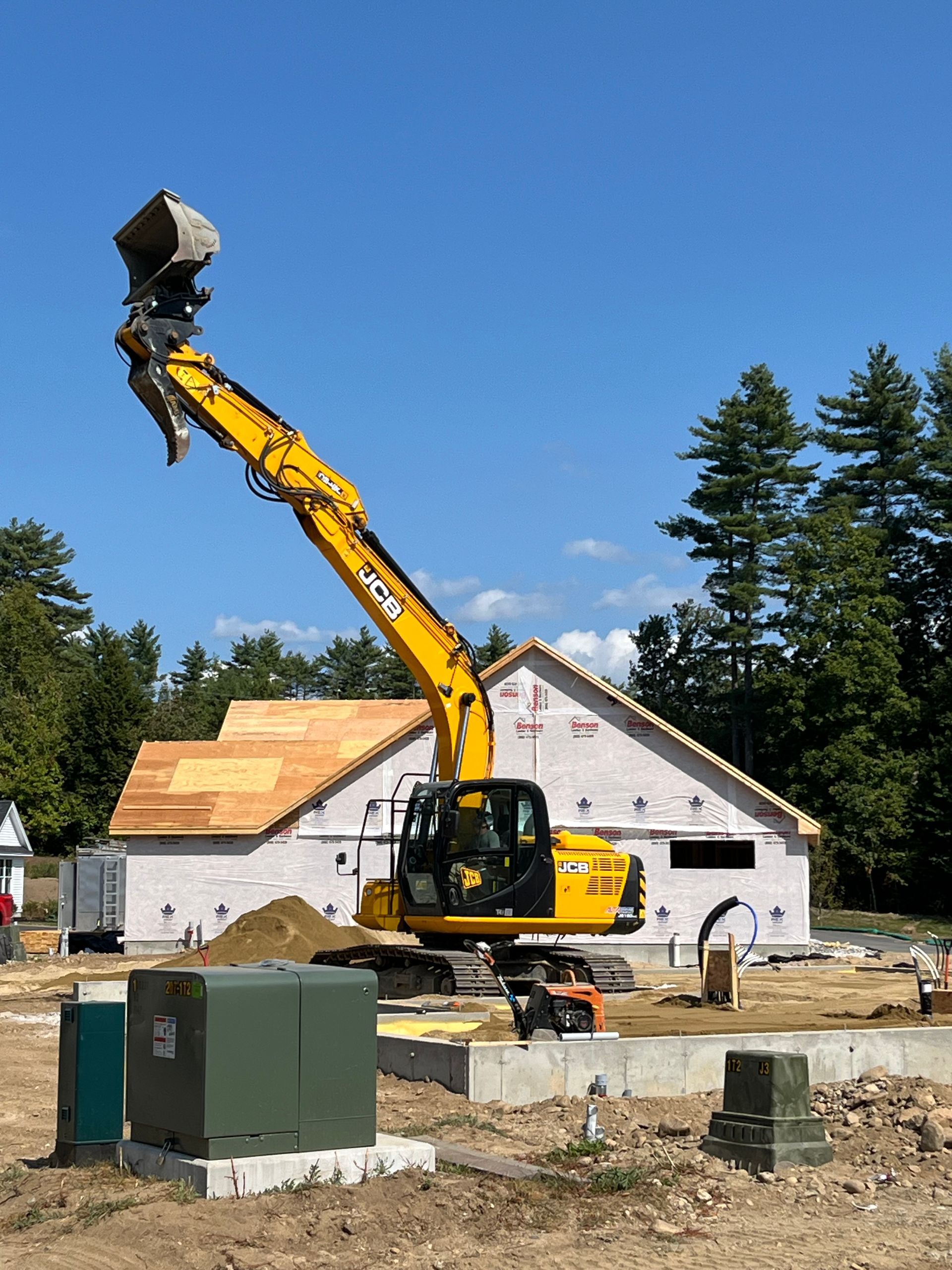 A yellow JCB excavator with an extended arm sits on a construction site in front of a house under construction.
