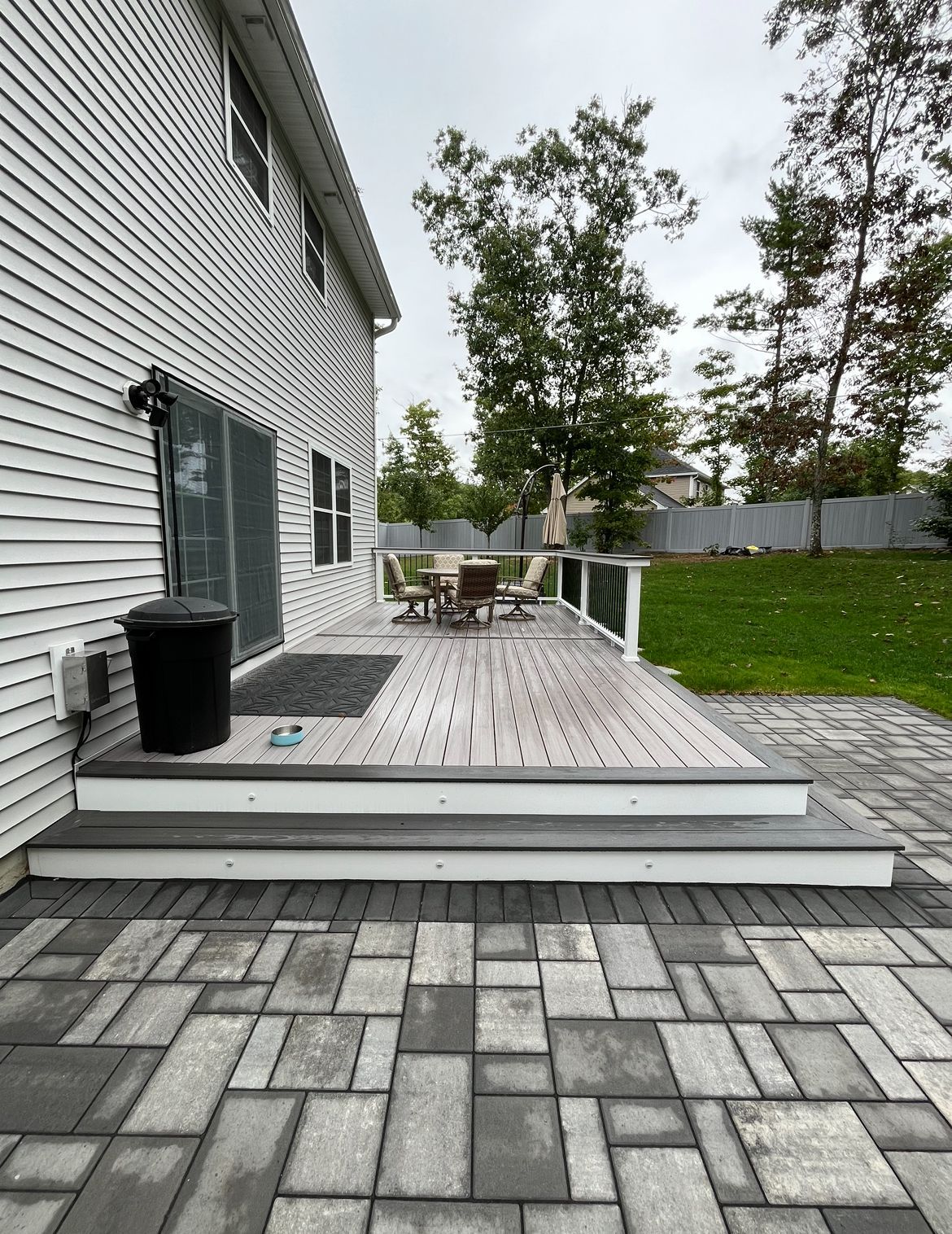 Gray deck with two steps leads from a house to a large gray paver patio, with a green lawn and trees in the background.