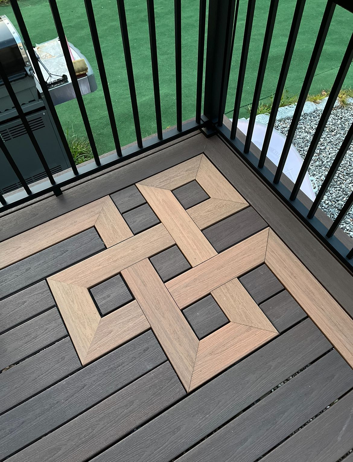 A light wood-grain Celtic knot inlay set into a dark gray composite deck, bordered by black metal railings.