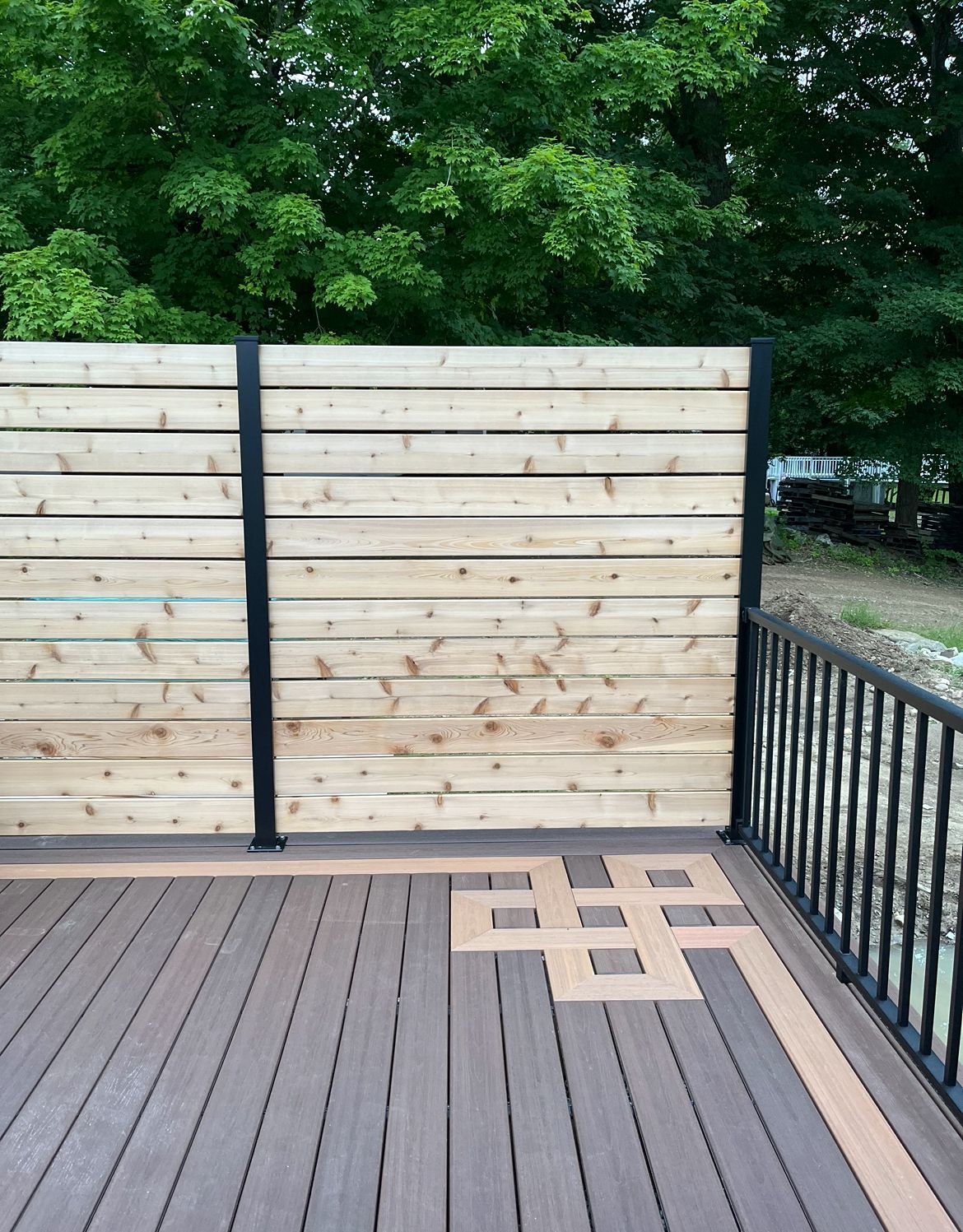 A deck with brown composite boards, a geometric border pattern, black metal railings, and a horizontal wood privacy fence.