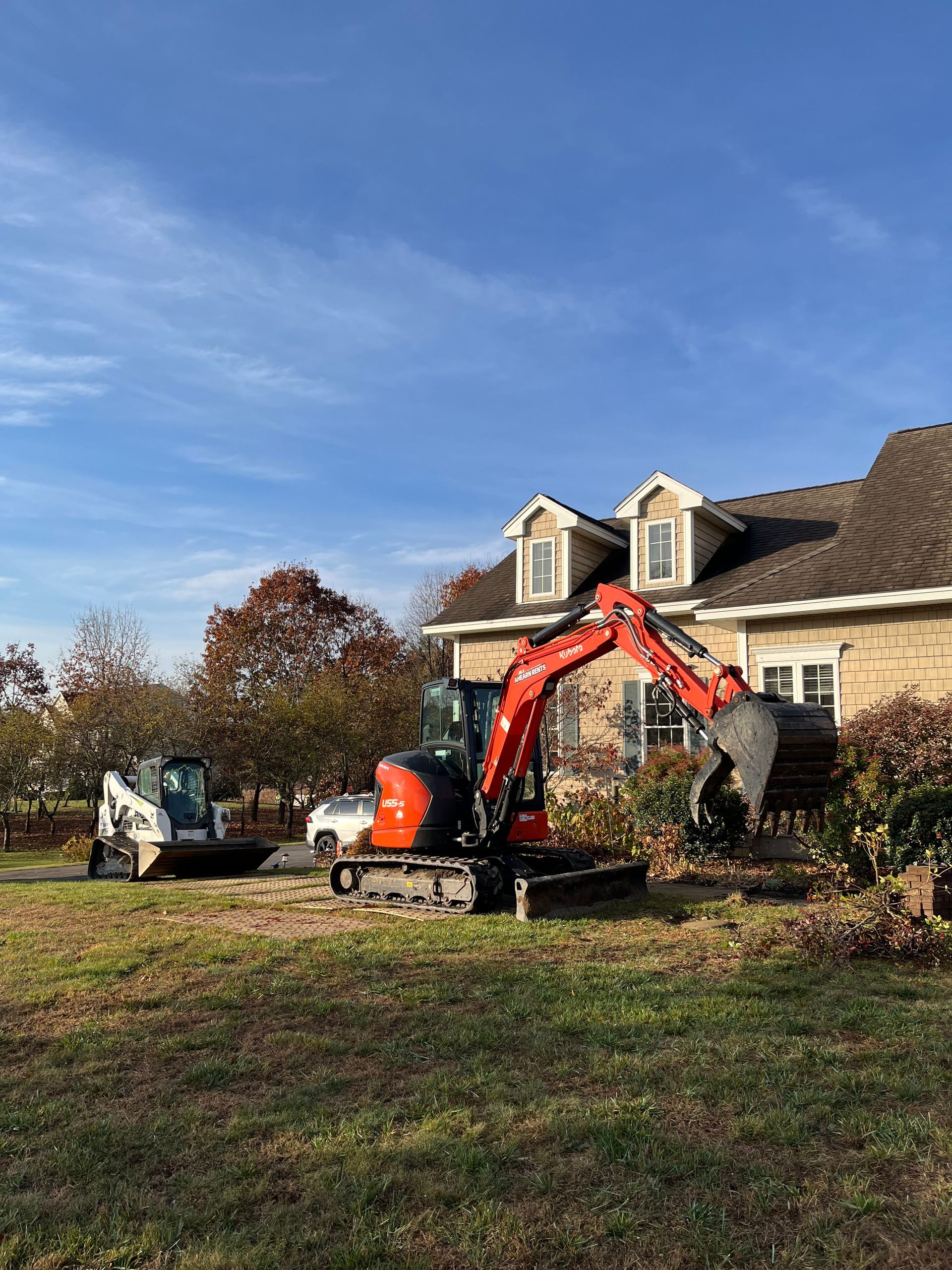 An orange excavator and a white skid steer parked on a grassy lawn in front of a house on a sunny day.