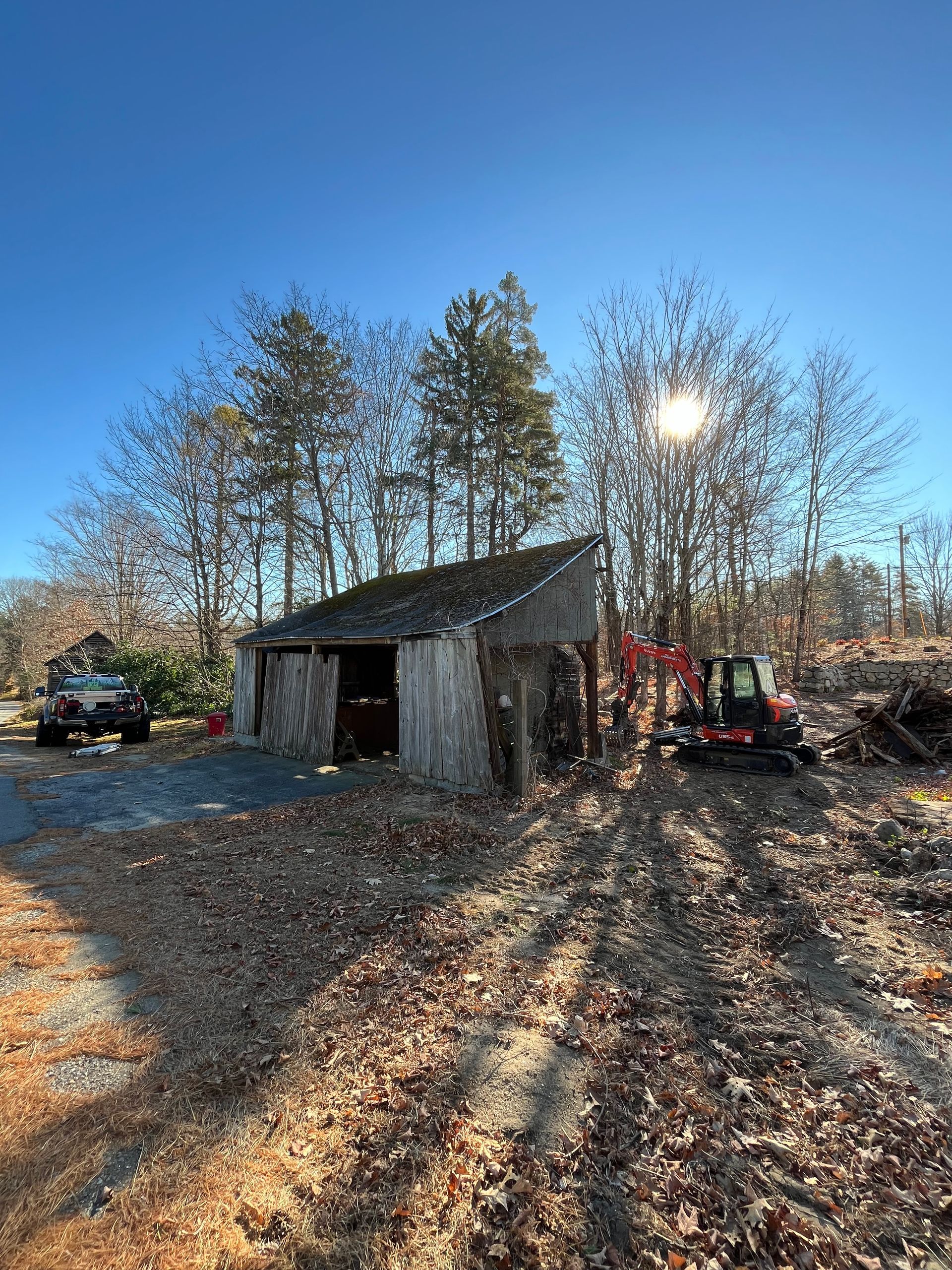 A sunlit rural yard featuring a weathered shed, a parked truck, and an orange mini-excavator near bare trees.