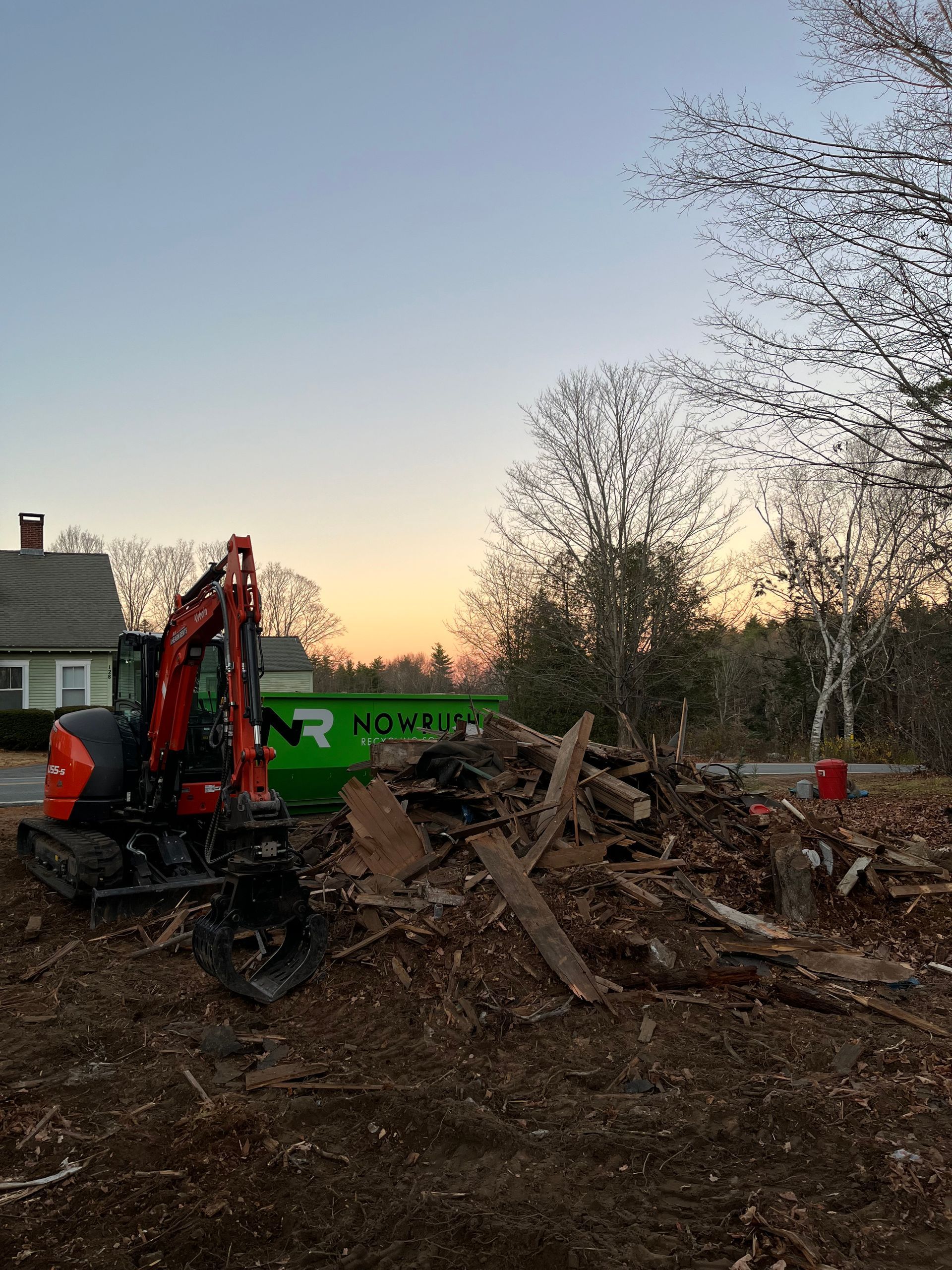 An orange excavator sits next to a green dumpster at a debris-filled demolition site at sunset.