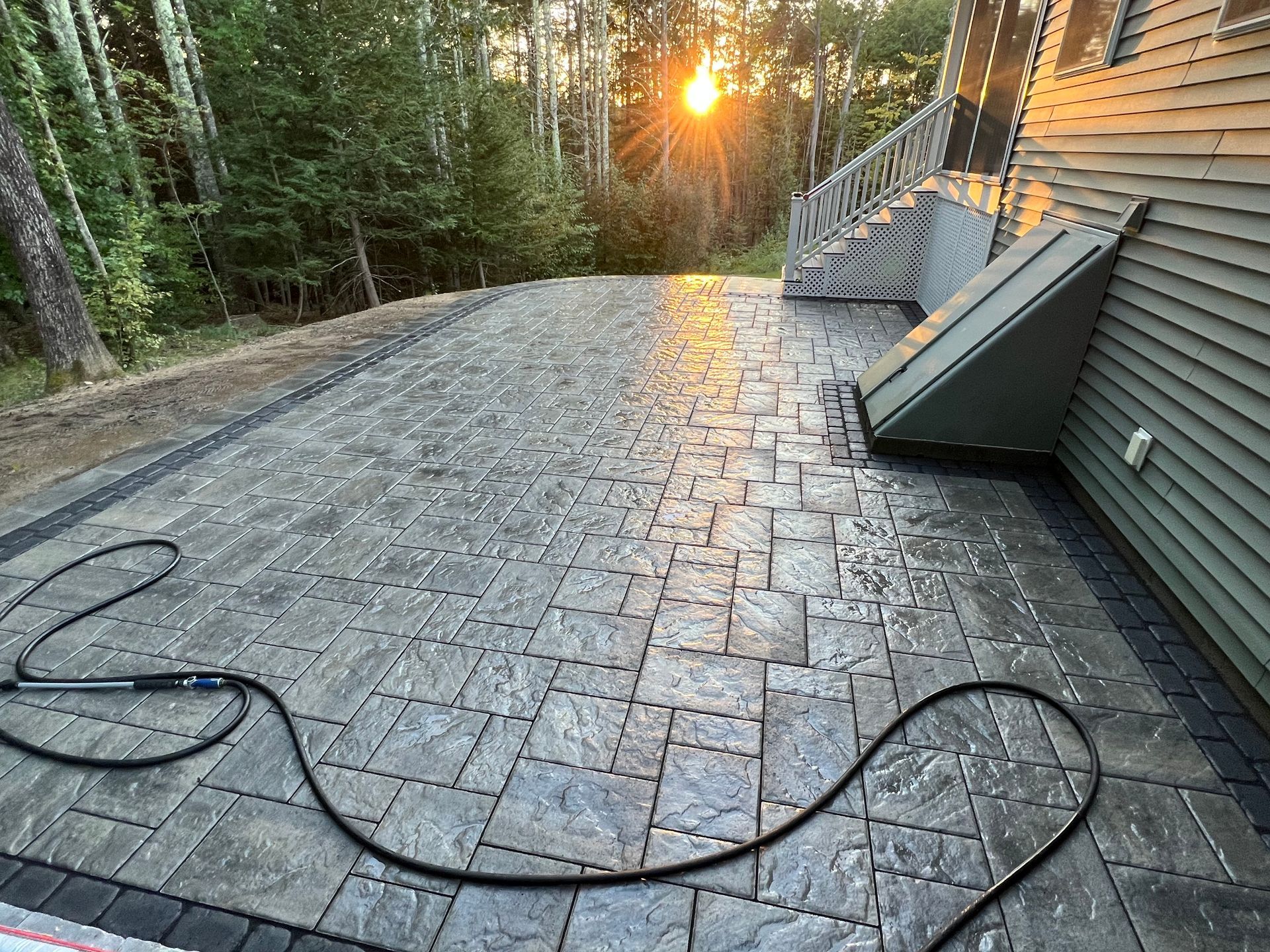 A freshly installed gray stone paver patio sits beside a house with a basement bulkhead, lit by a golden sunset.