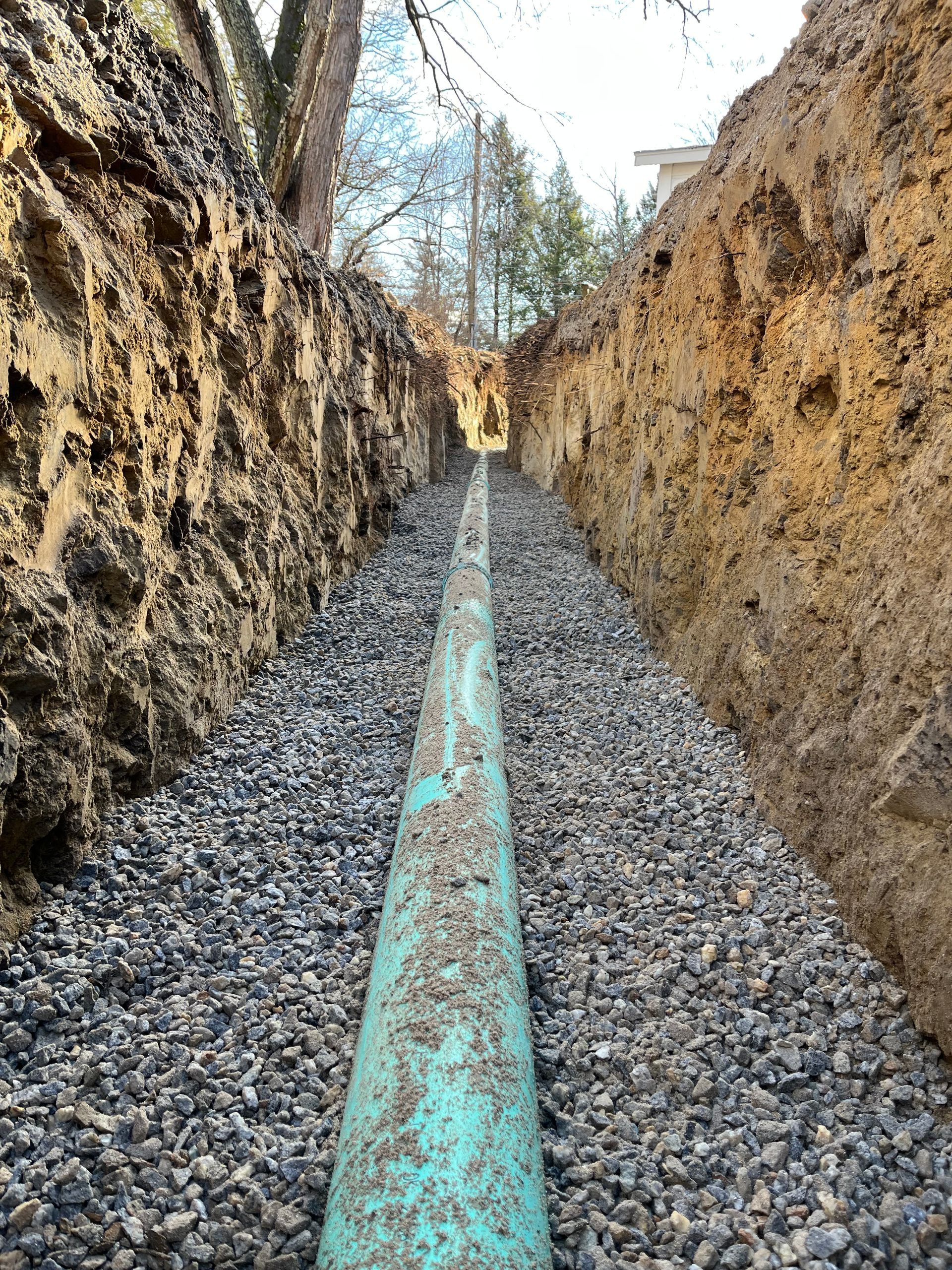 A green pipe lies in a gravel-filled trench between two dirt walls.