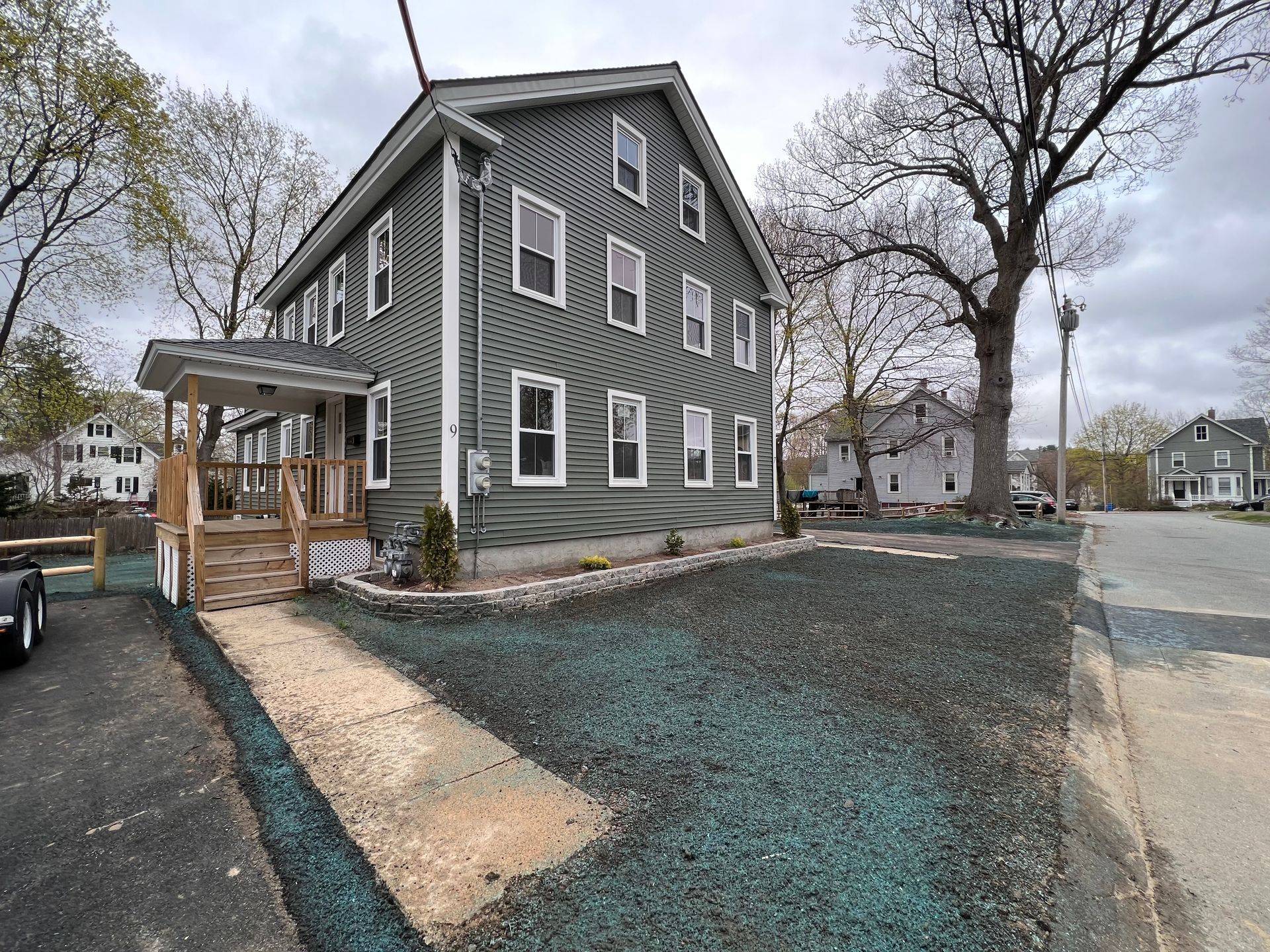 A three-story, grey, sided house with a front porch, a newly graveled yard, and a concrete path, set in a residential area.