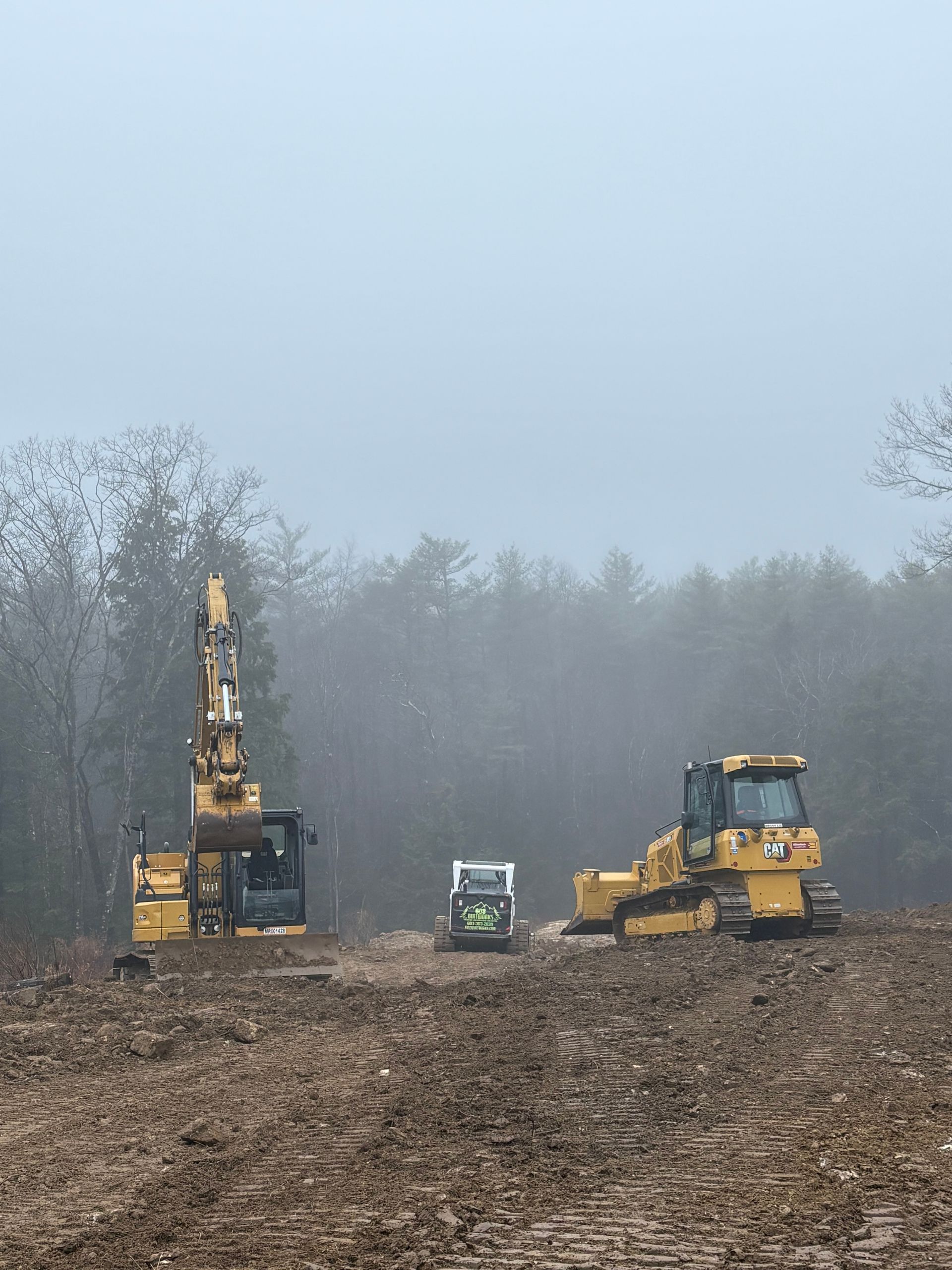 Three yellow construction vehicles work on a muddy, dirt-covered clearing under a foggy, overcast sky.