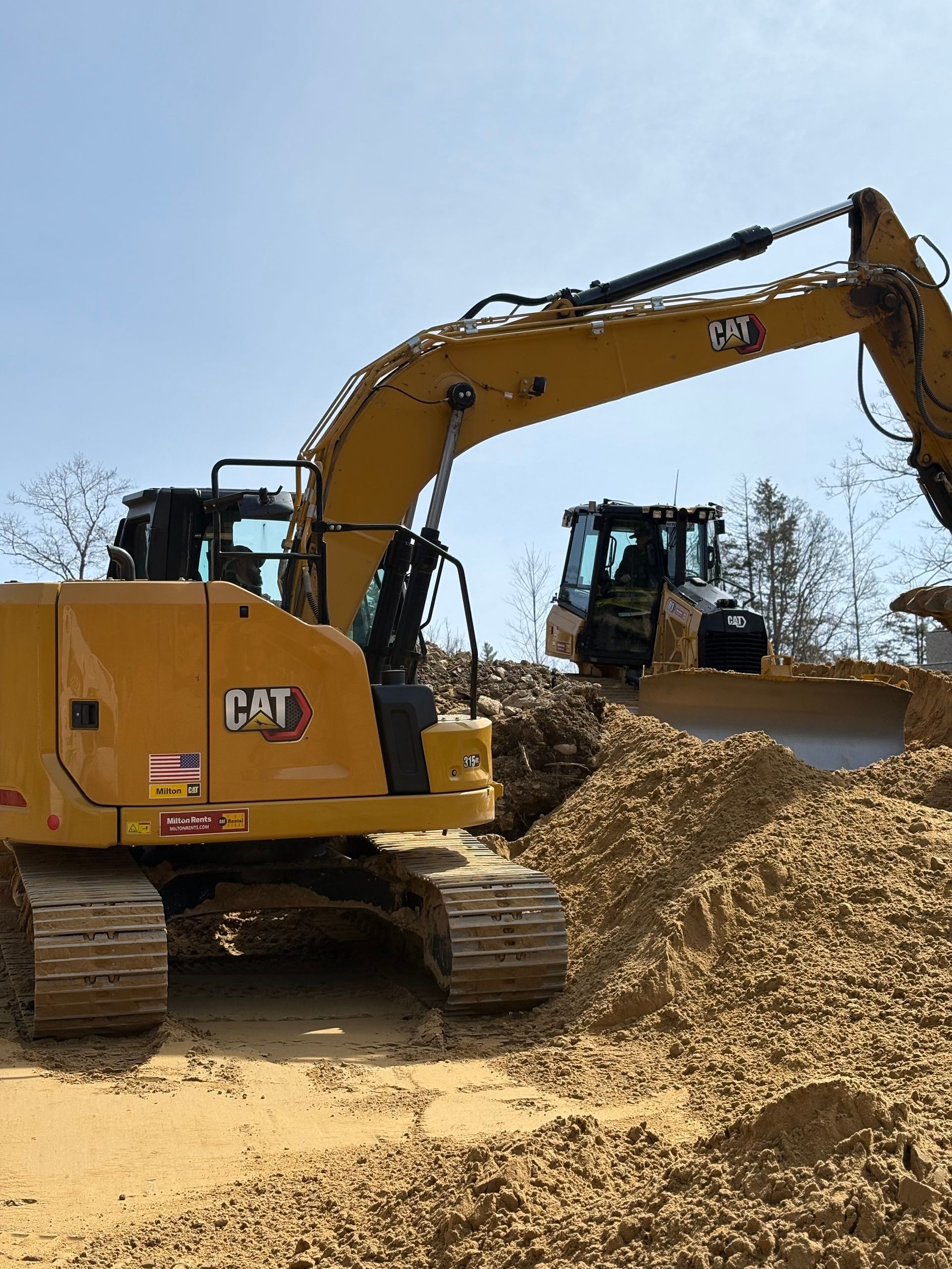 Two yellow Caterpillar excavators work on a dirt construction site under a bright sky.