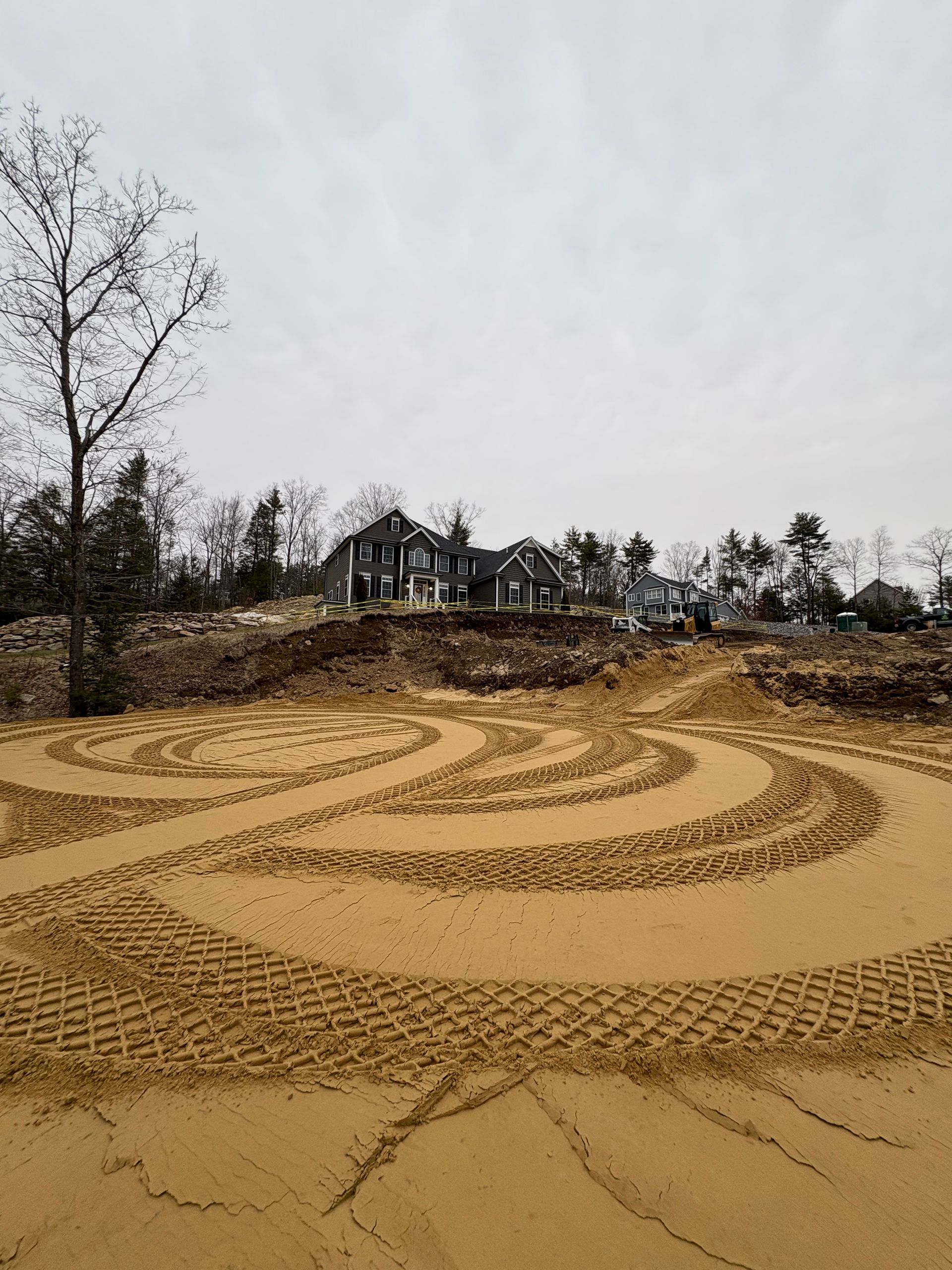 Large, swirling tire tracks left by construction machinery in a field of sand, with a residential house in the background.