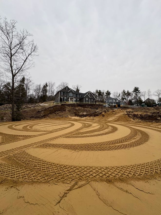 Large, swirling tire tracks left by construction machinery in a field of sand, with a residential house in the background.
