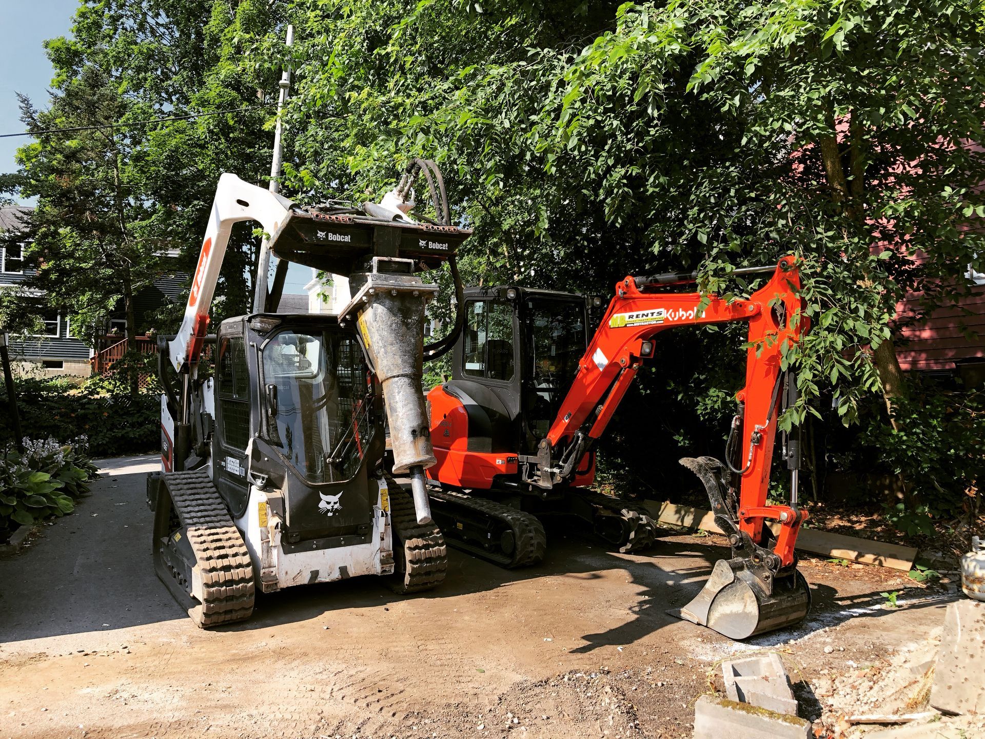 A white Bobcat skid-steer with a hydraulic breaker attachment parked next to an orange mini-excavator in a yard.