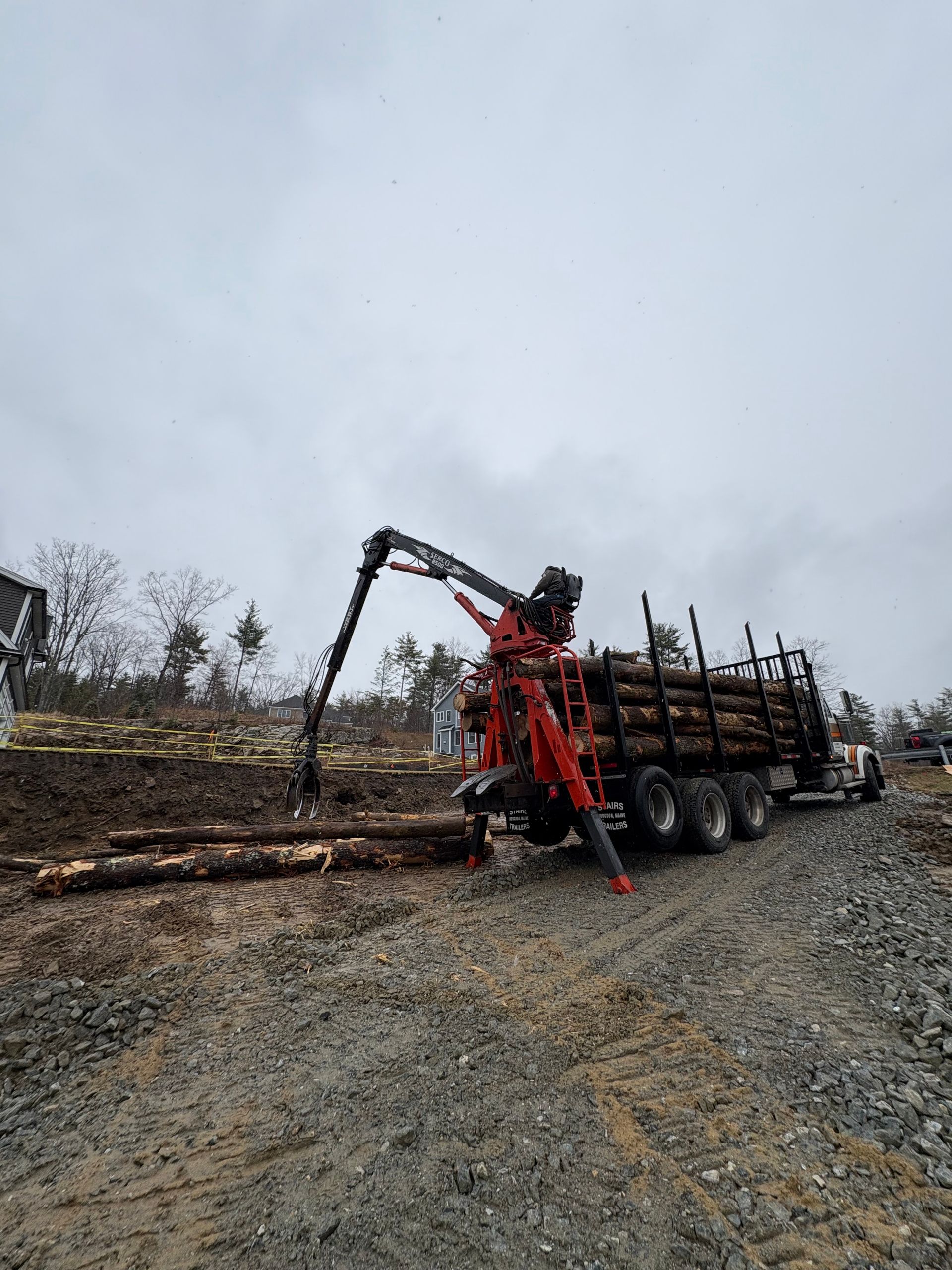 A red logging truck parked on a gravel road uses its hydraulic crane to load felled logs onto its trailer.