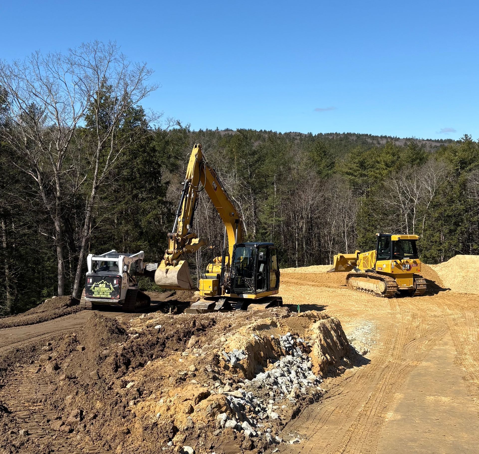 A yellow excavator, a white skid steer, and a yellow bulldozer clear land on a sunny day with trees in the background.