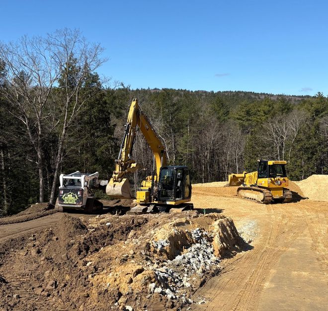 A yellow excavator, a white skid steer, and a yellow bulldozer clear land on a sunny day with trees in the background.