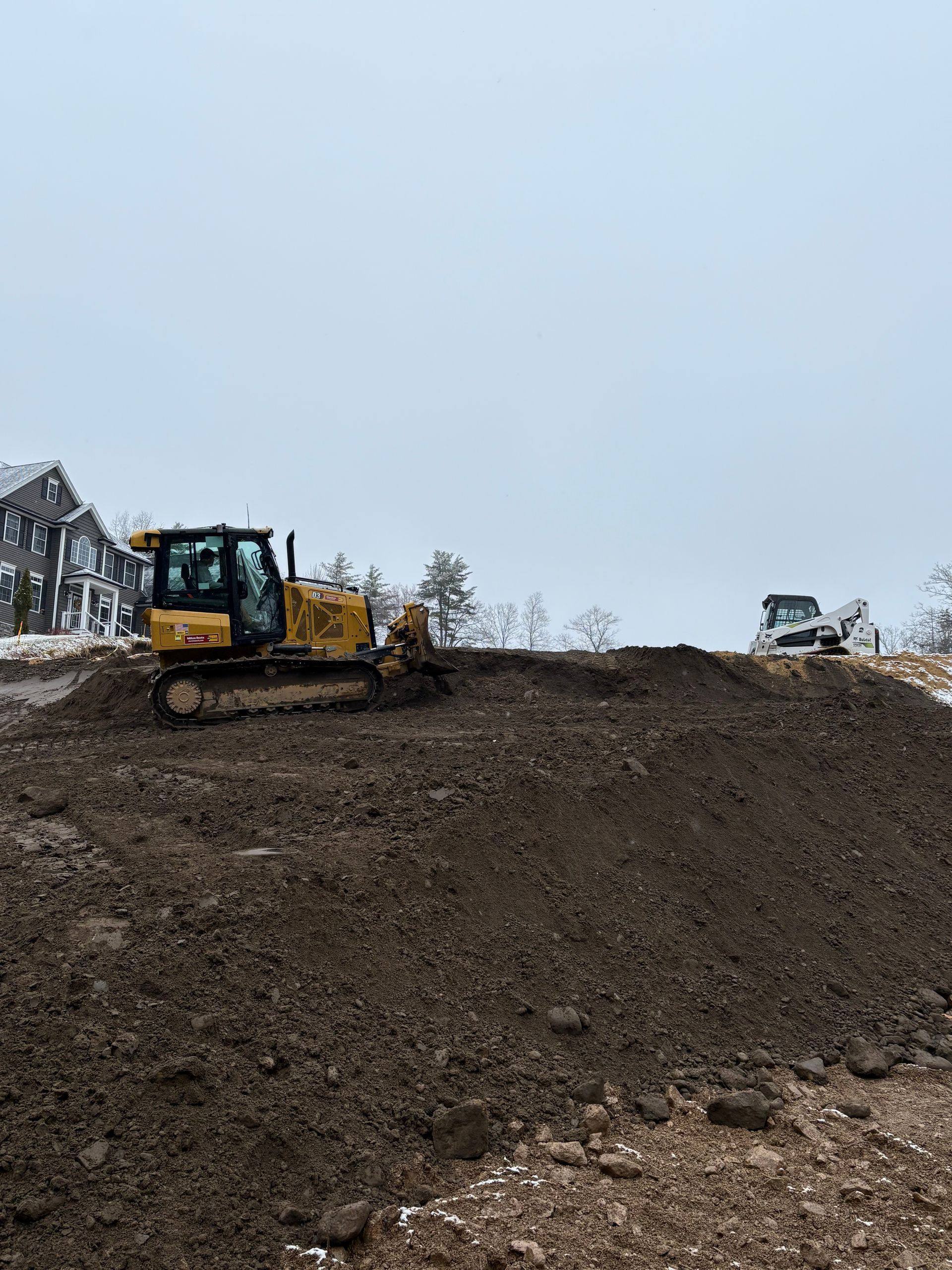 A yellow bulldozer sits atop a large mound of dark soil at a construction site under a gray, overcast sky.