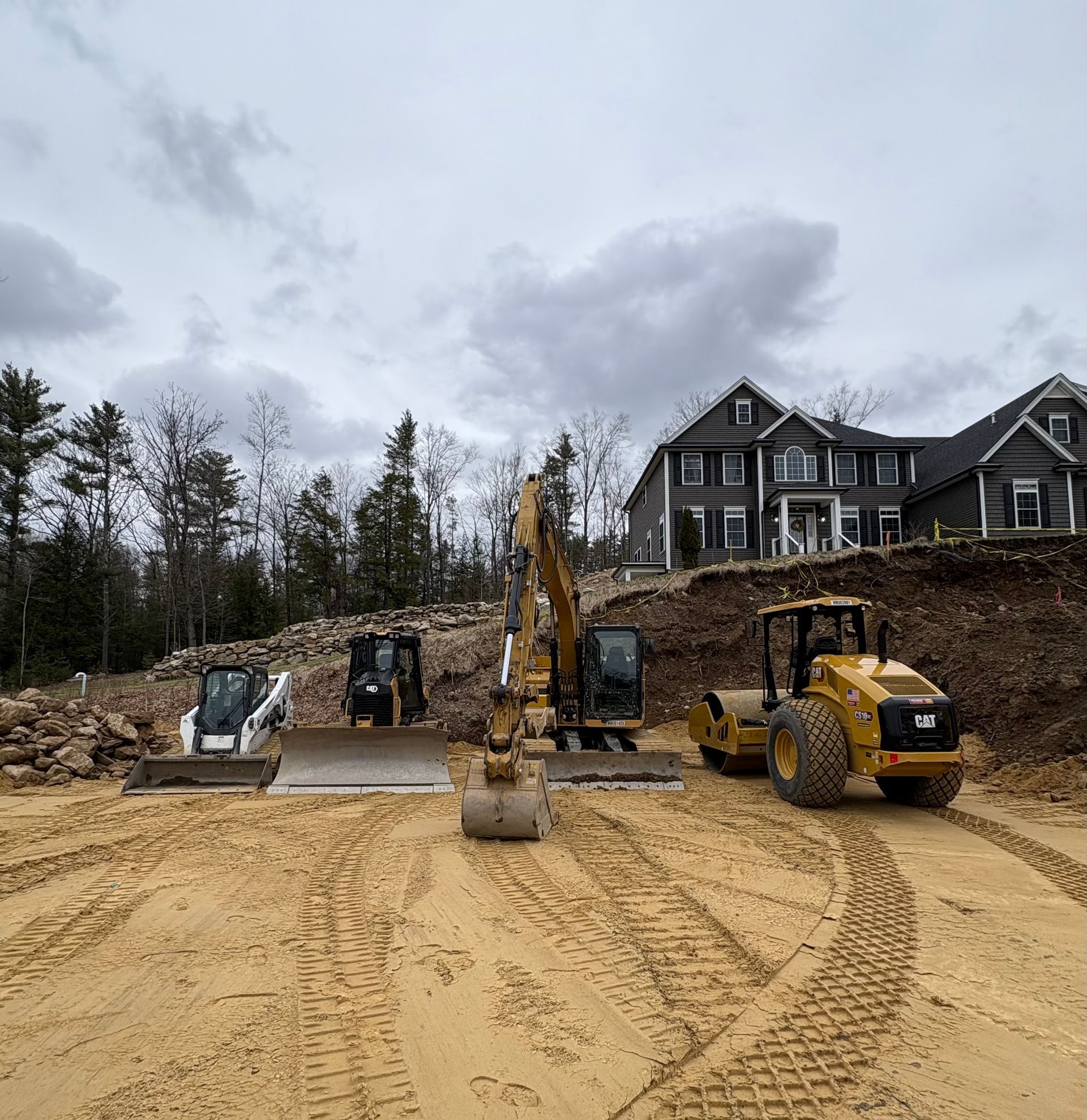 Construction equipment, including a roller and an excavator, parked on a dirt lot in front of a house under construction.