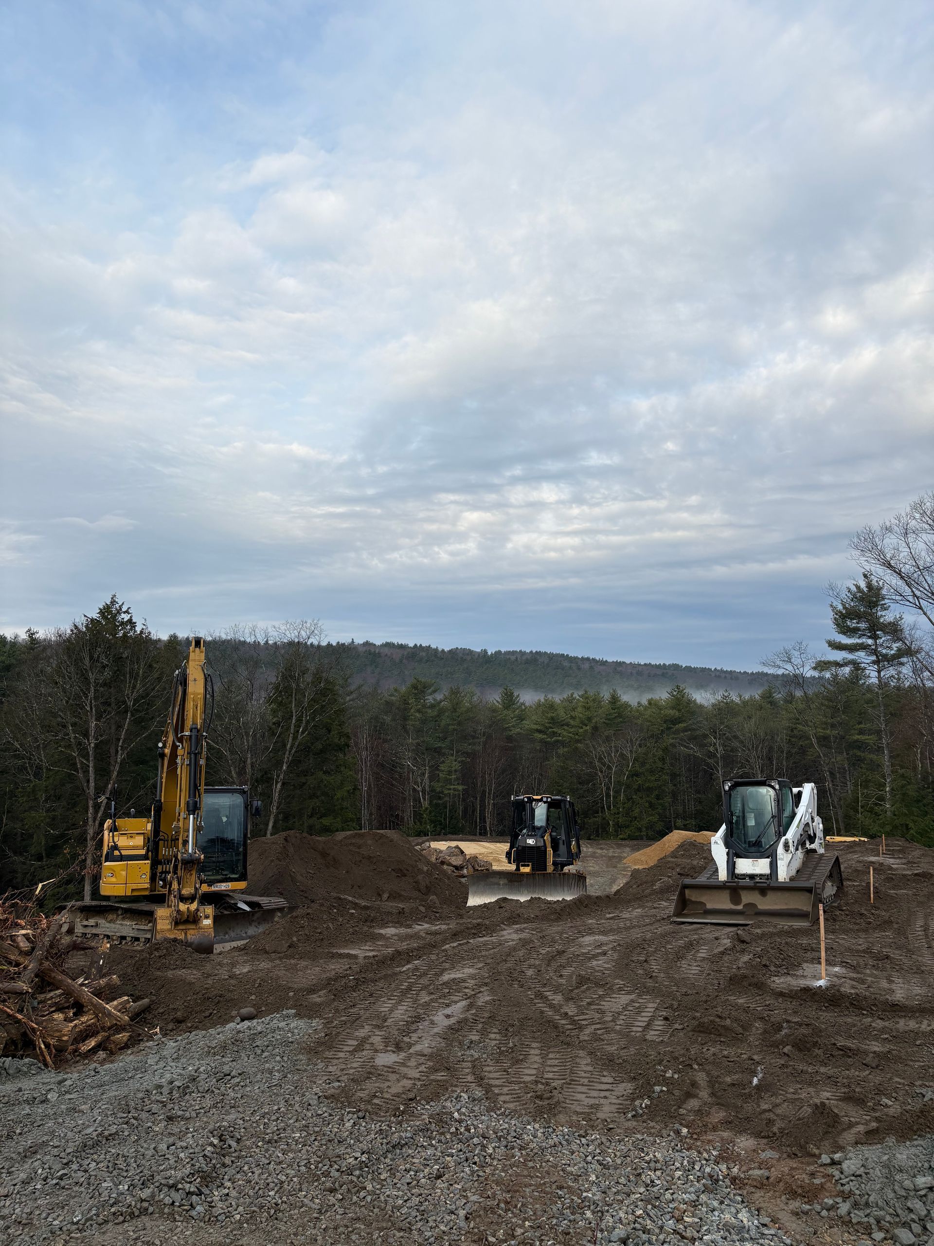 Construction site with heavy machinery, including an excavator and a loader, clearing land near a forest at dusk.