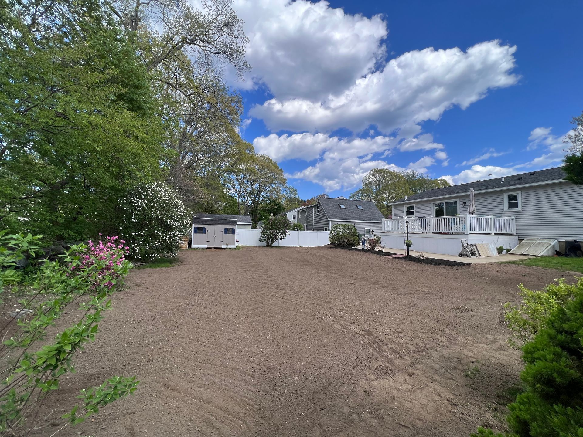 A large, freshly leveled backyard area with brown soil, a white storage shed, and a house with a deck under a cloudy sky.