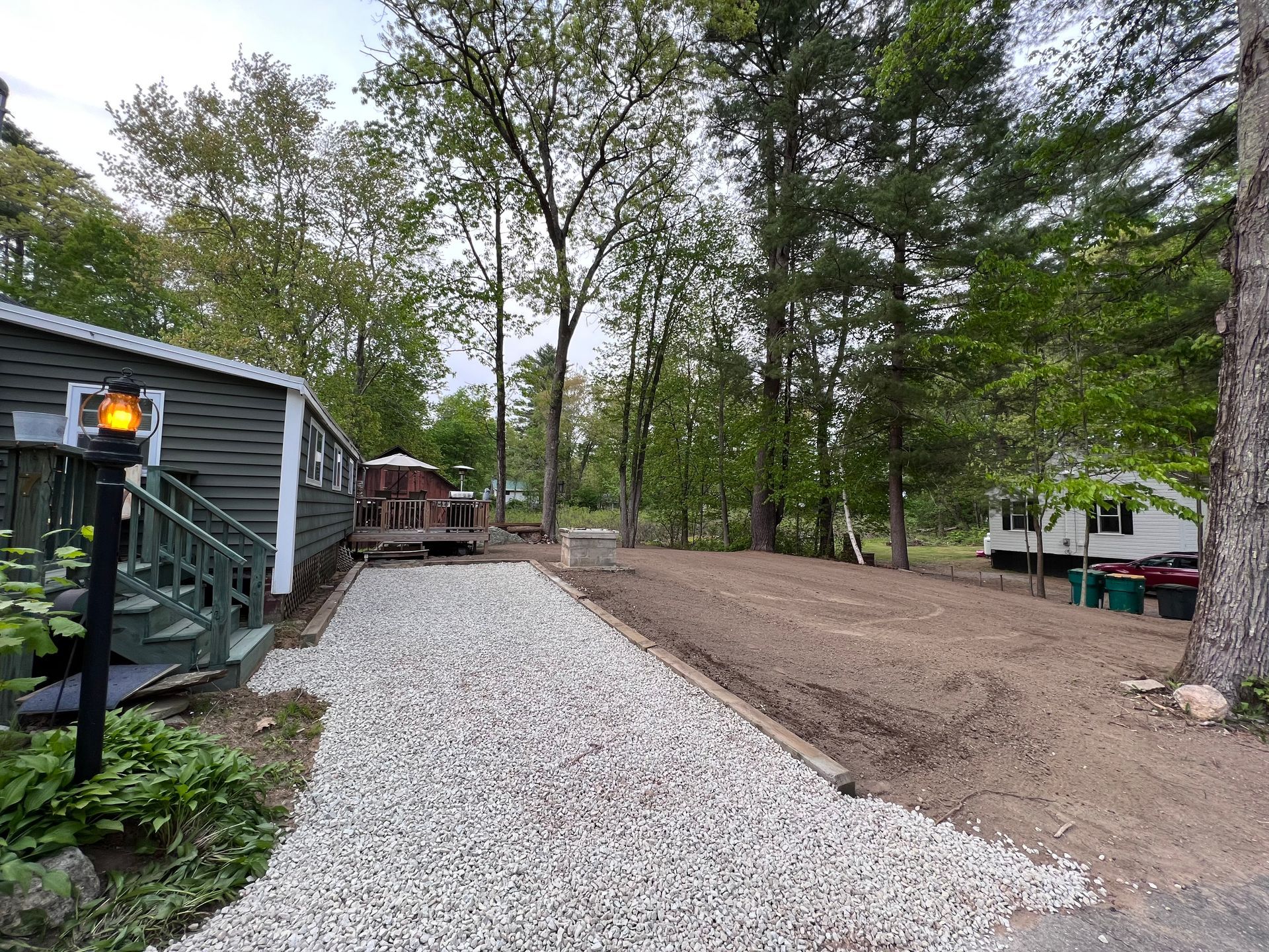 A grey building with a stone-covered path leading to a wooded yard with an RV in the distance on a sunny day.