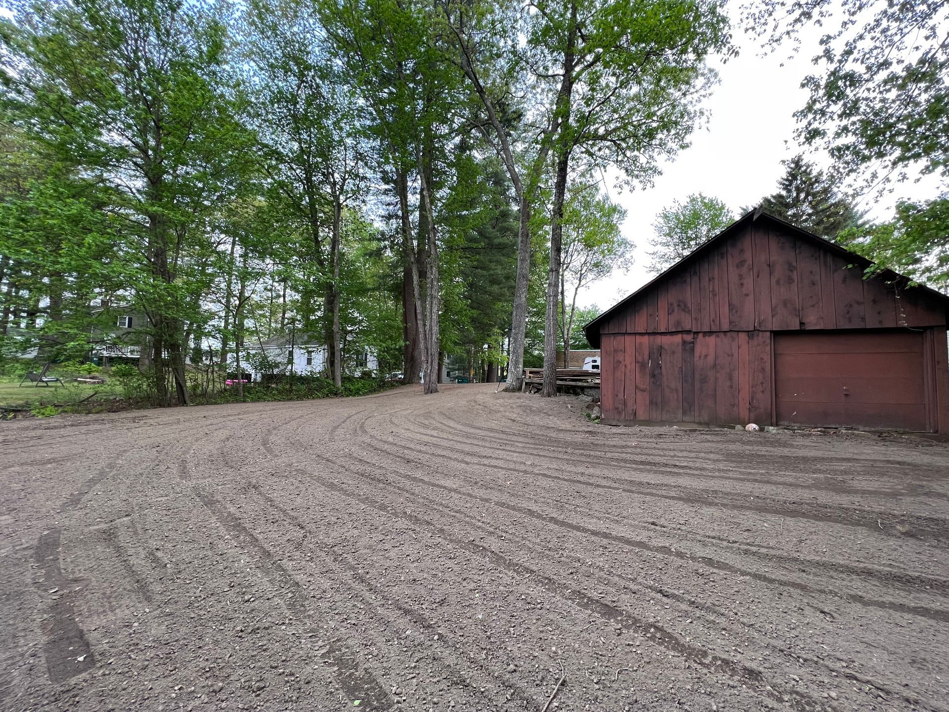 A gravel driveway leading to a red wooden barn nestled among lush green trees on a bright day.