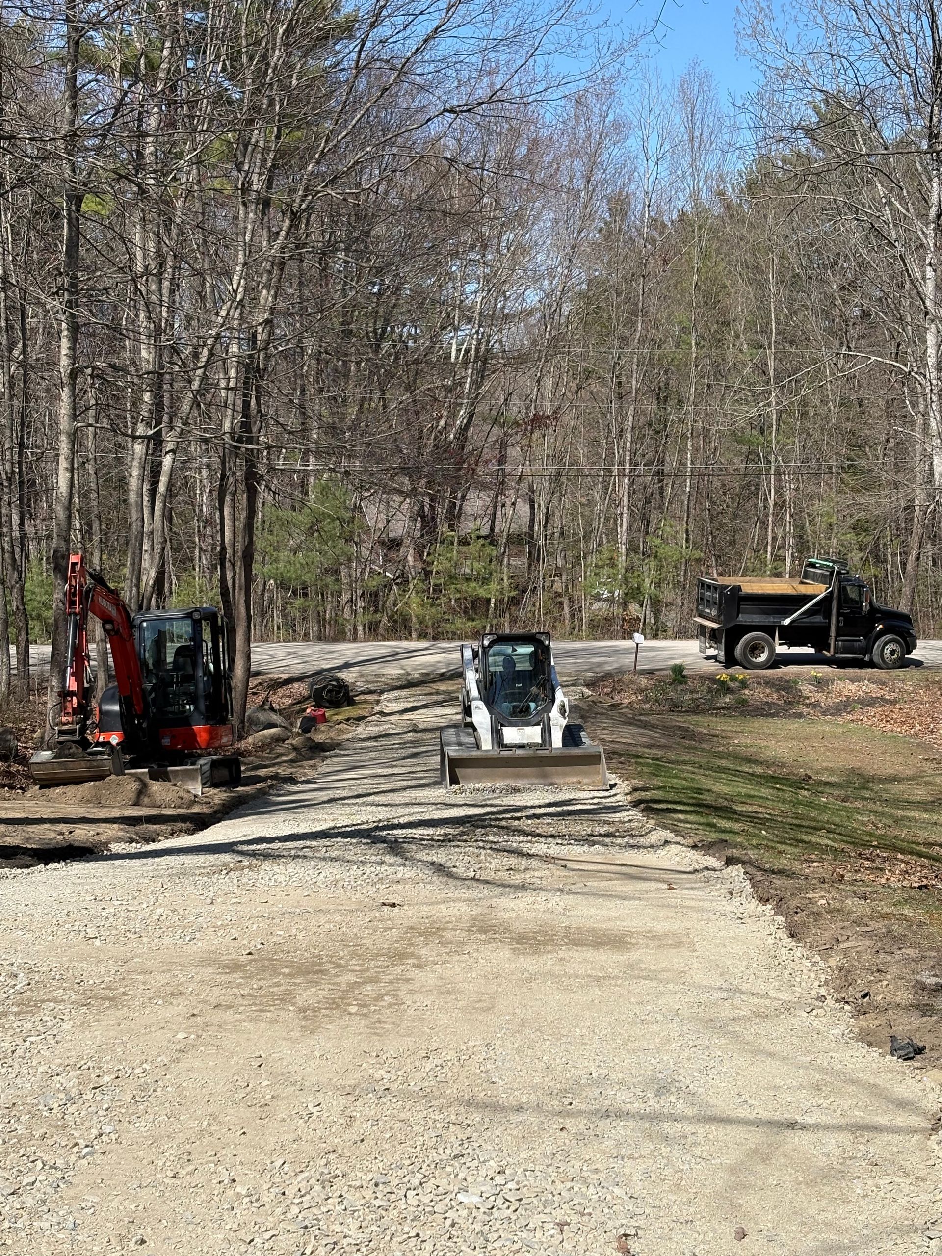 Construction site in a wooded area with an excavator, a skid steer, and a black dump truck parked on a gravel driveway.