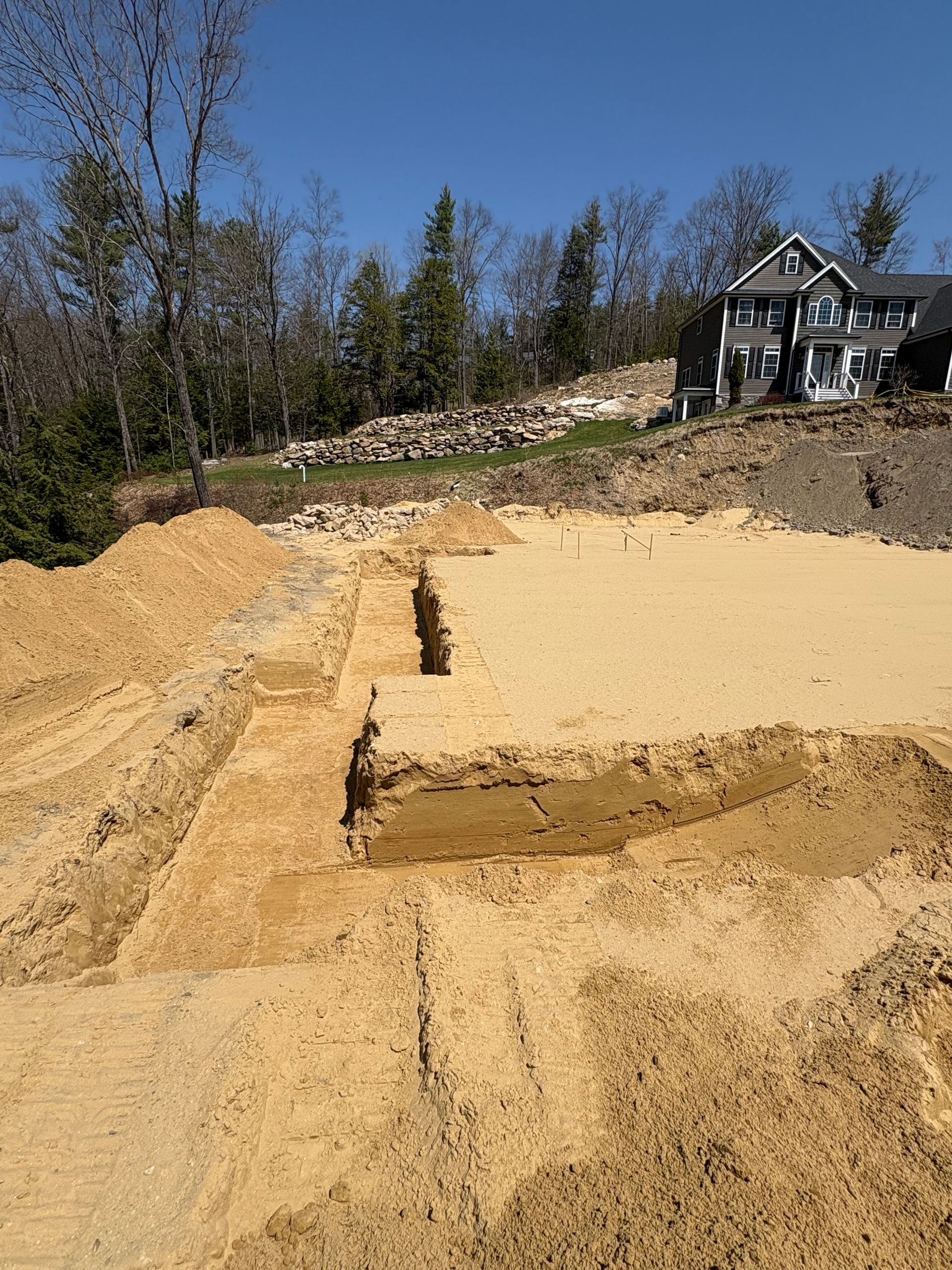 An excavation site with cleared tan soil, trench foundations, and a house in the background under a clear blue sky.