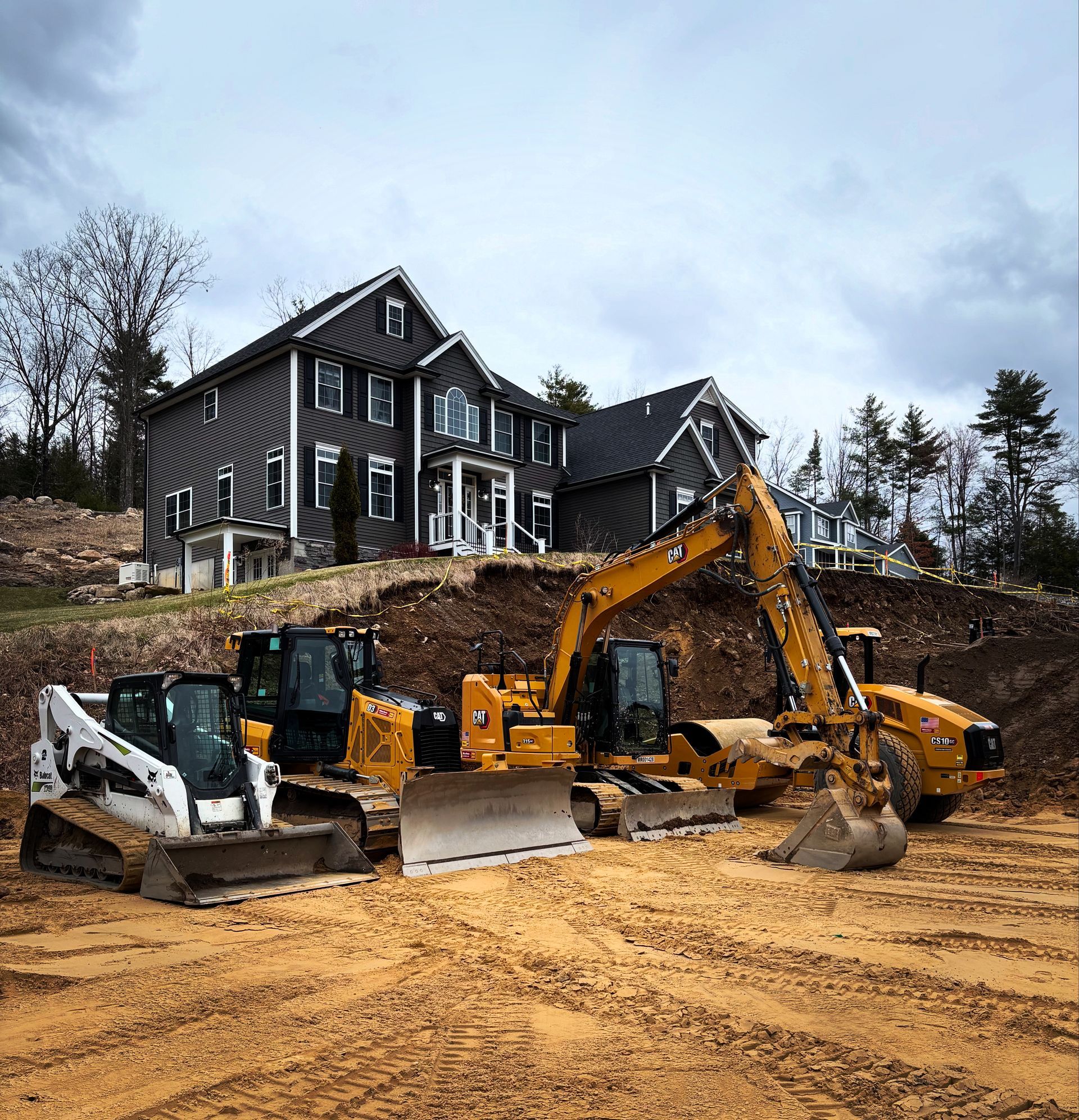 Three pieces of heavy construction equipment parked on a dirt lot in front of a large, dark-sided multi-story house.