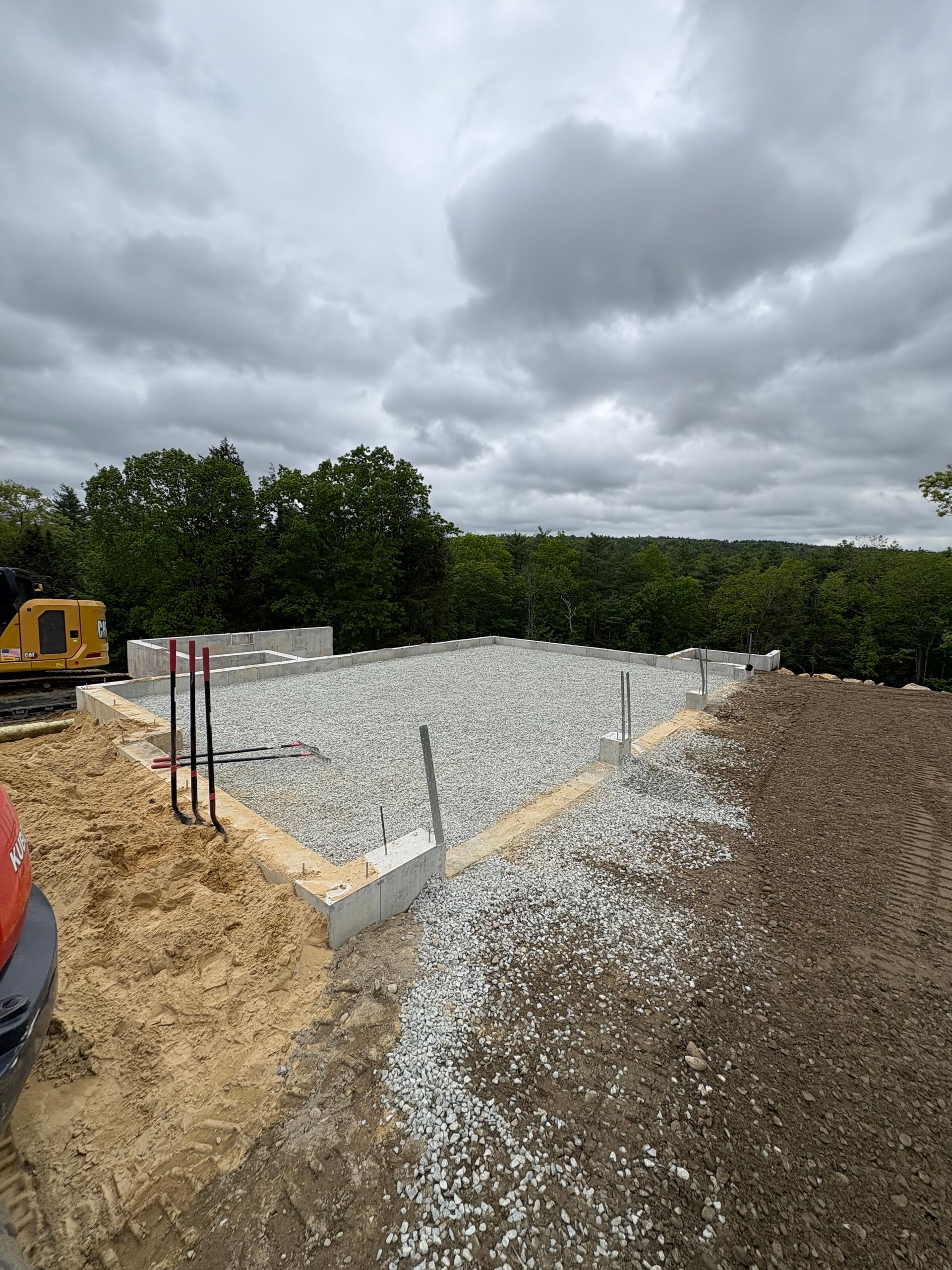 A concrete foundation for a building filled with gravel, surrounded by dirt and trees under a cloudy sky.
