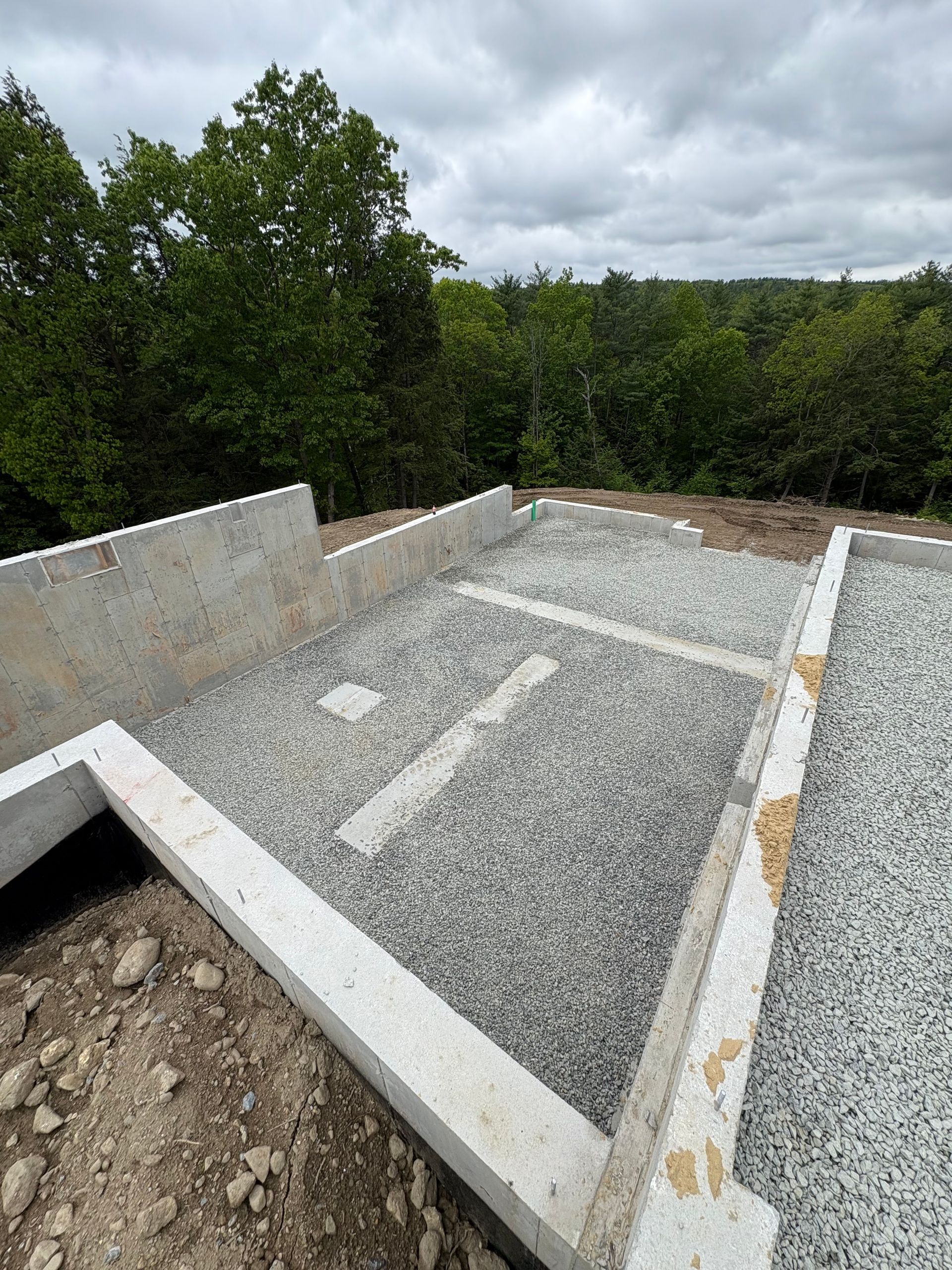 A construction site with concrete foundation walls and a gravel-filled floor, surrounded by a forest under a cloudy sky.