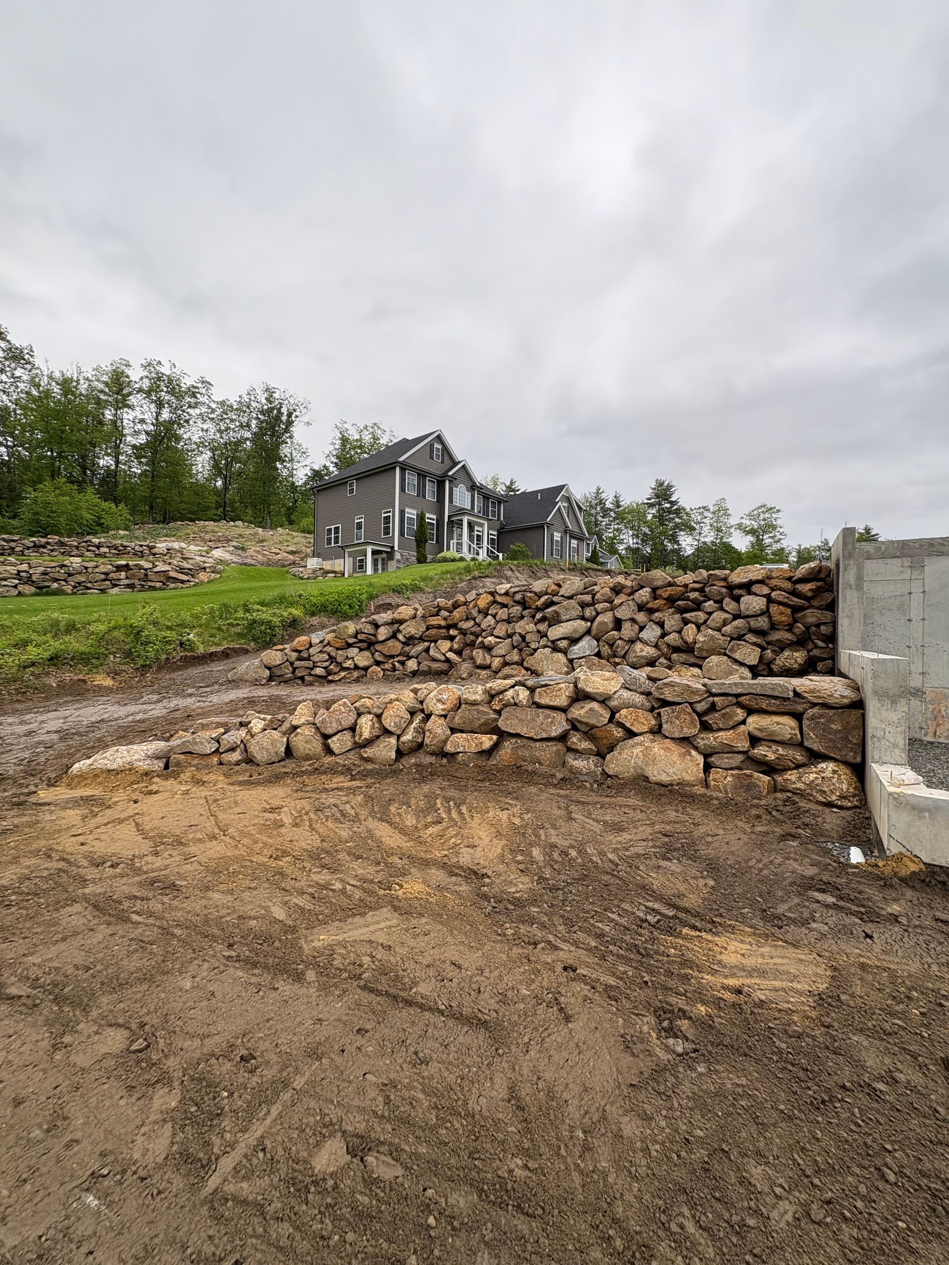 Stacked stone retaining walls leading up to modern houses on a hillside under a cloudy sky.