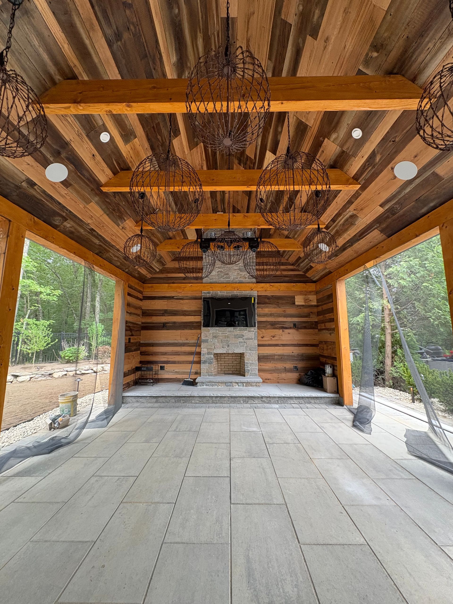 An outdoor stone patio under a rustic wooden pavilion featuring a central stone fireplace and warm overhead lighting.