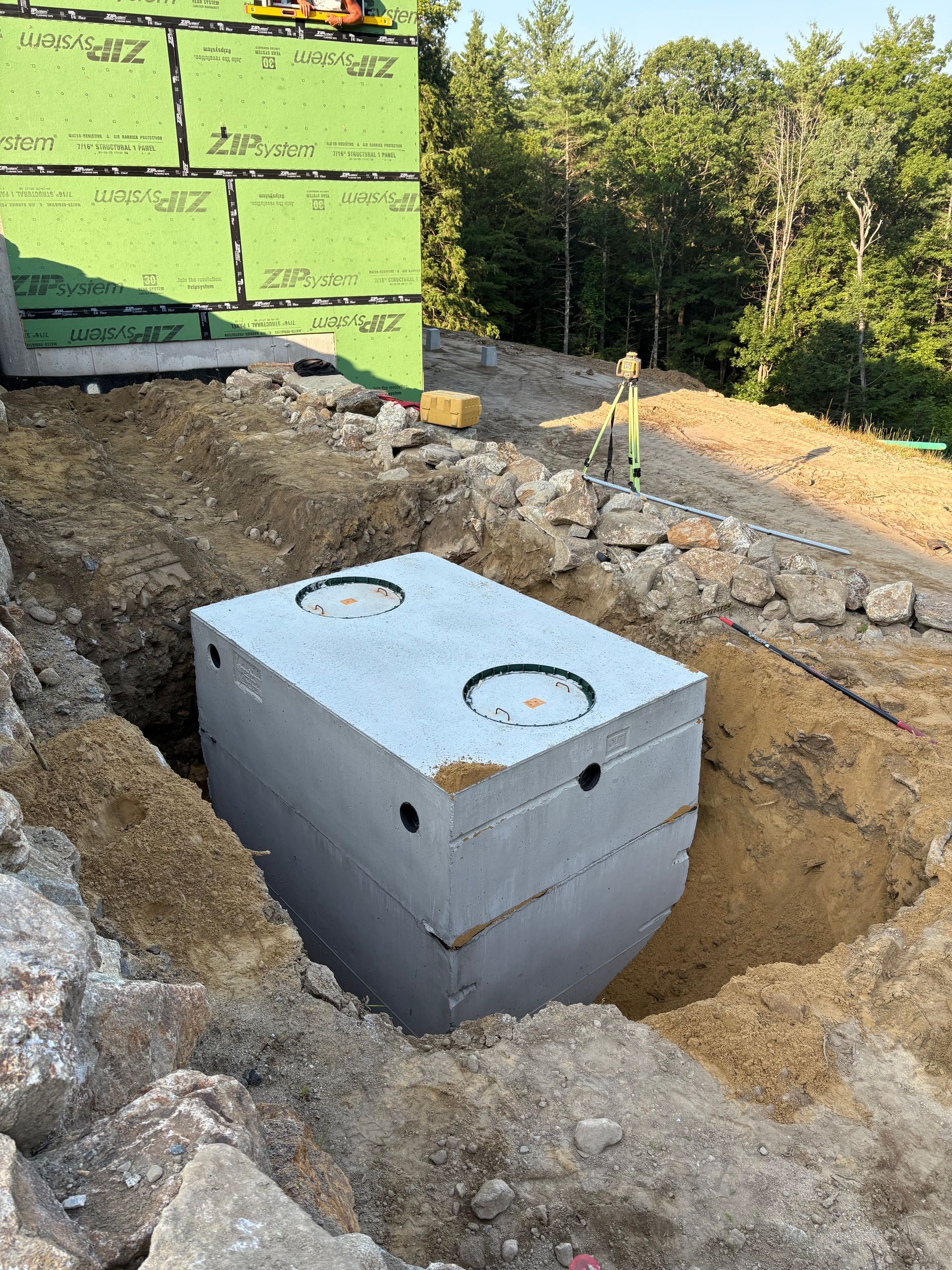 A concrete septic tank sits in an excavated trench next to a building under construction.