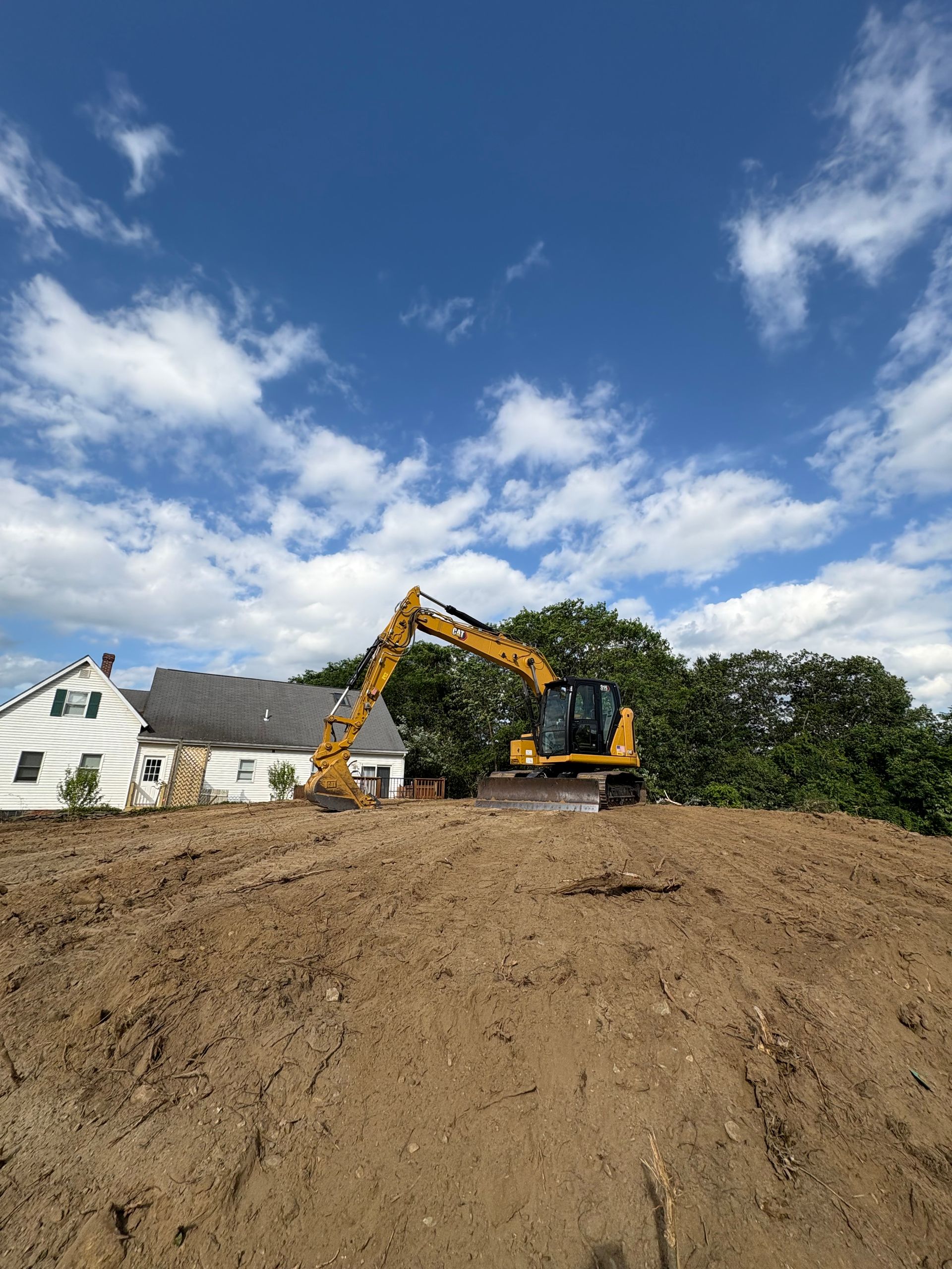 A yellow excavator sits atop a dirt mound near a white house against a blue, cloudy sky.