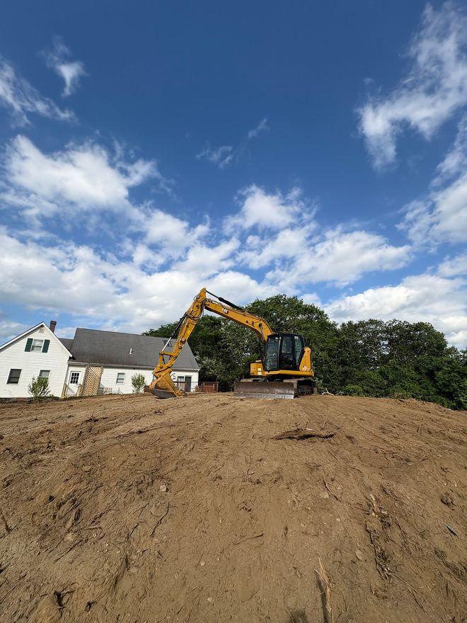 A yellow excavator sits atop a dirt mound near a white house against a blue, cloudy sky.