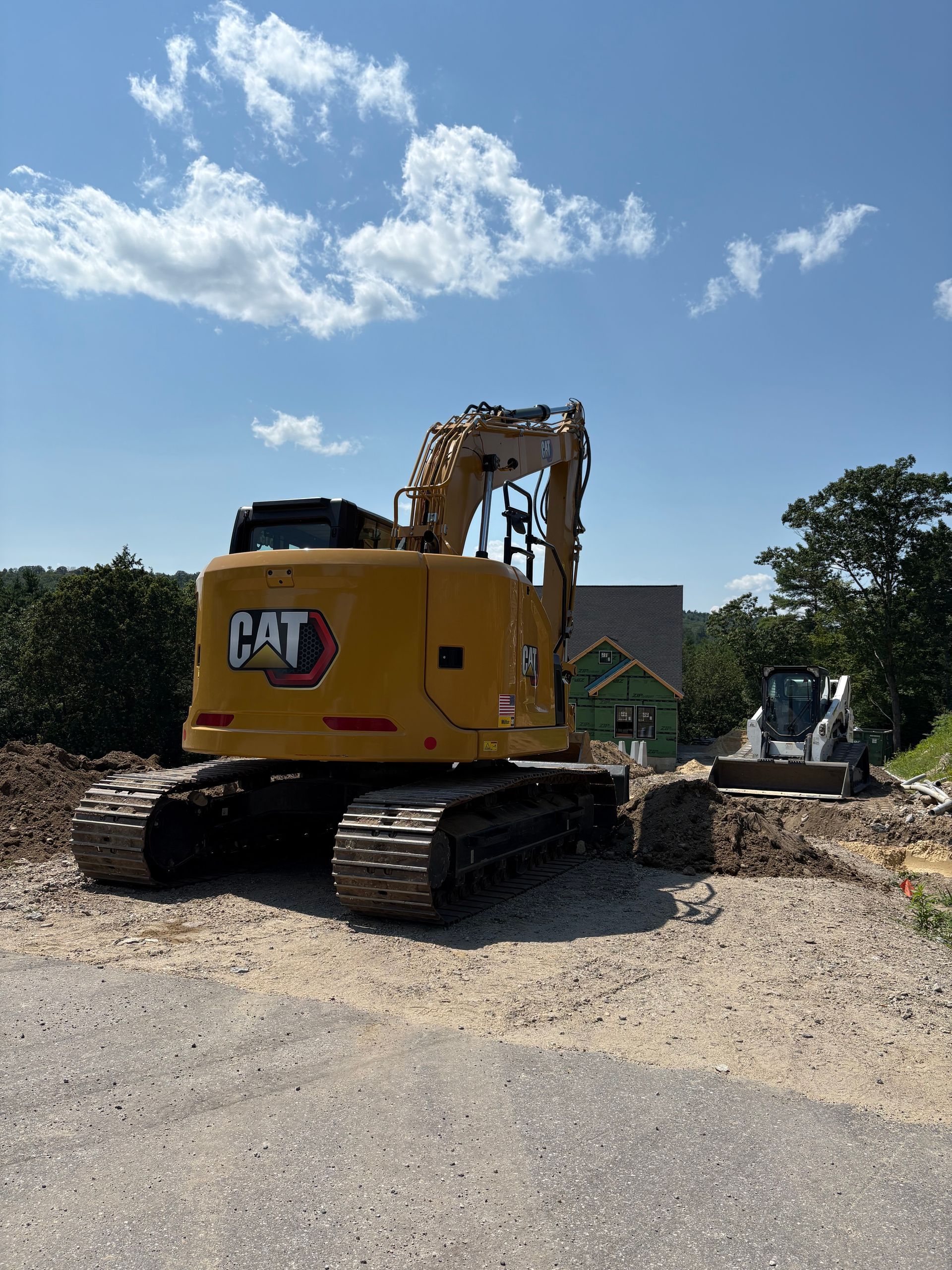 Yellow CAT excavator parked on a dirt lot at a construction site with a house frame in the background under a blue sky.