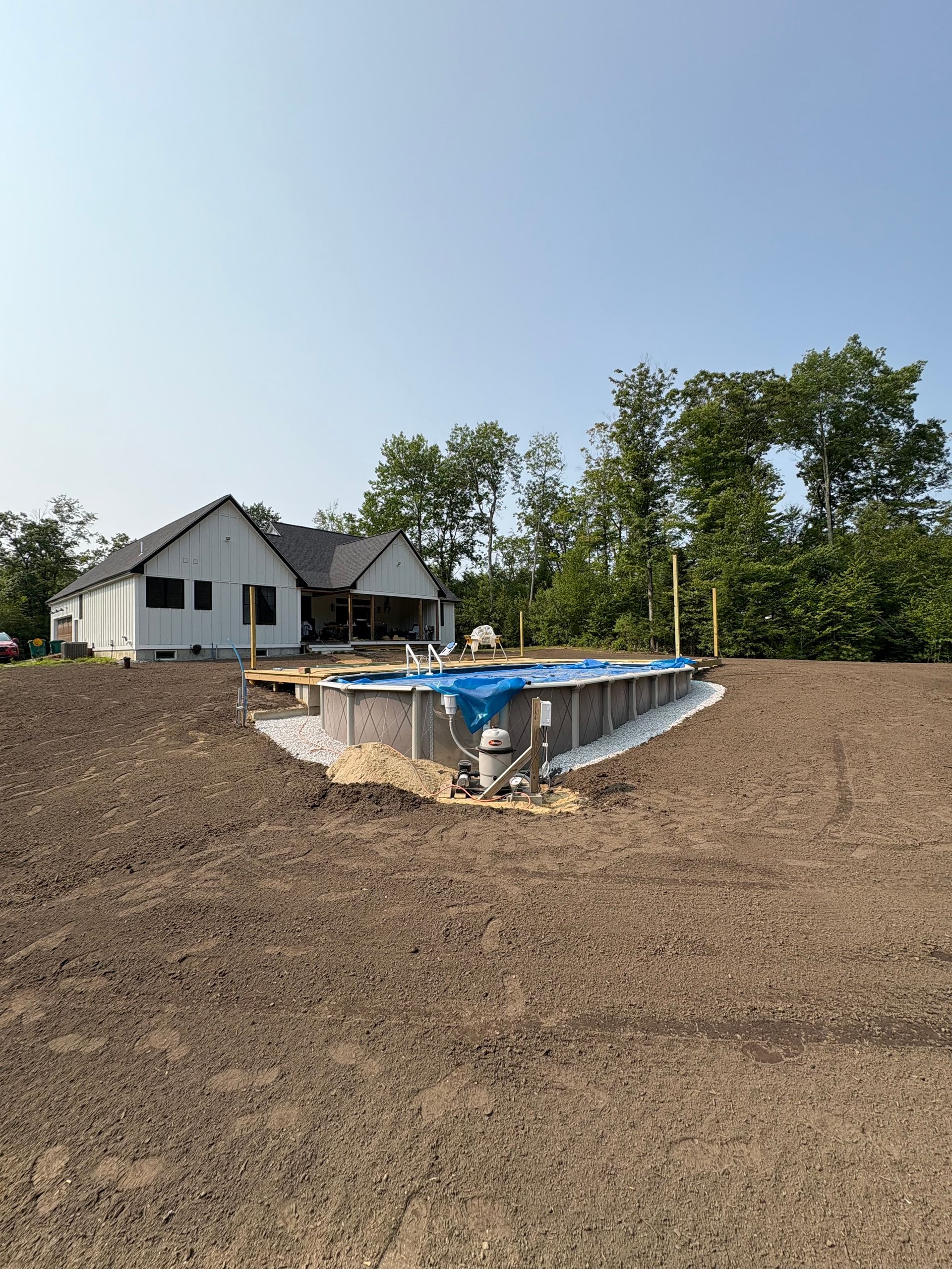 An above-ground pool sits in a large, dirt-covered yard in front of a white house with a dark roof on a sunny day.