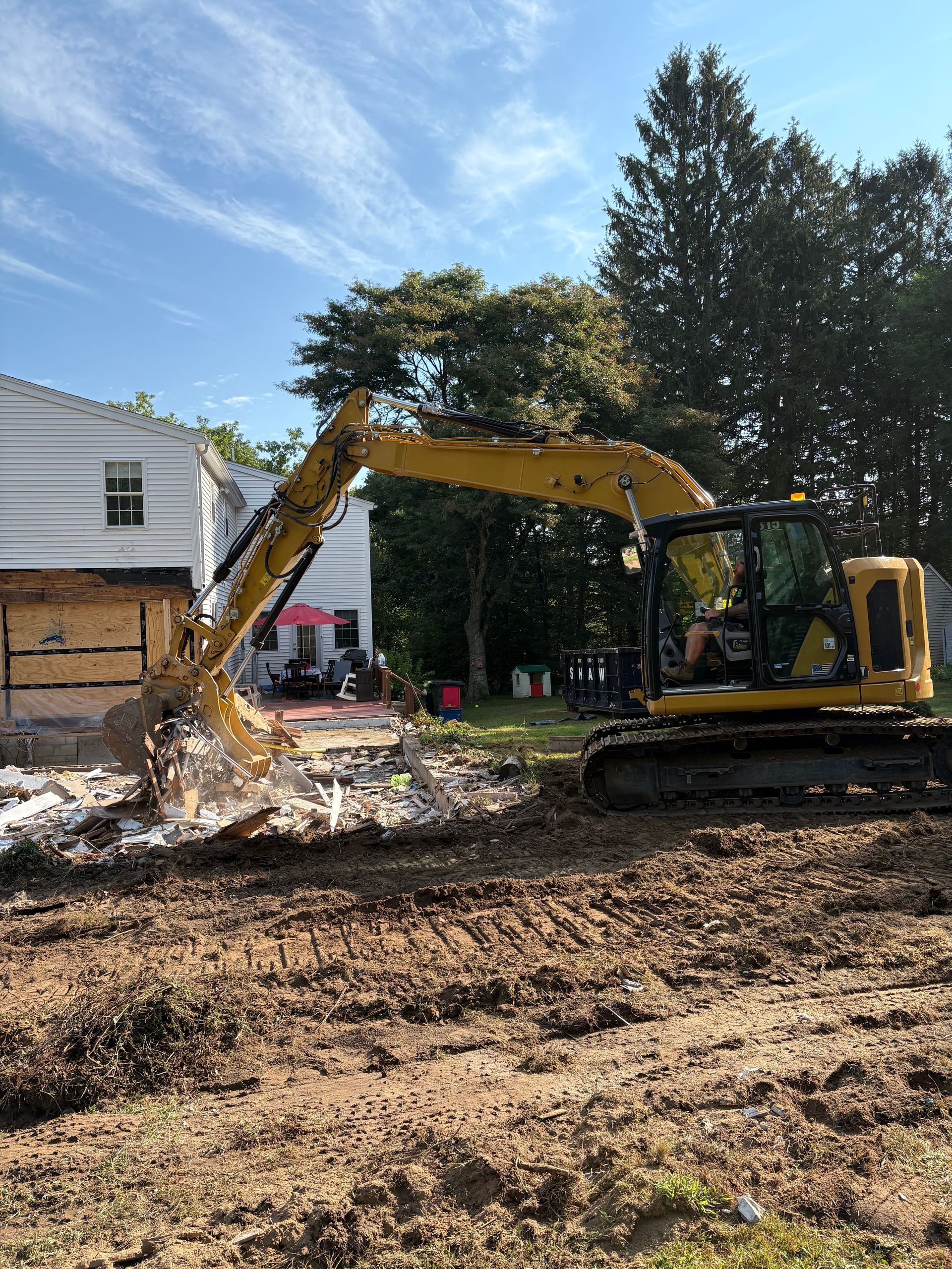 A yellow excavator demolishes a section of a white two-story house on a sunny day.