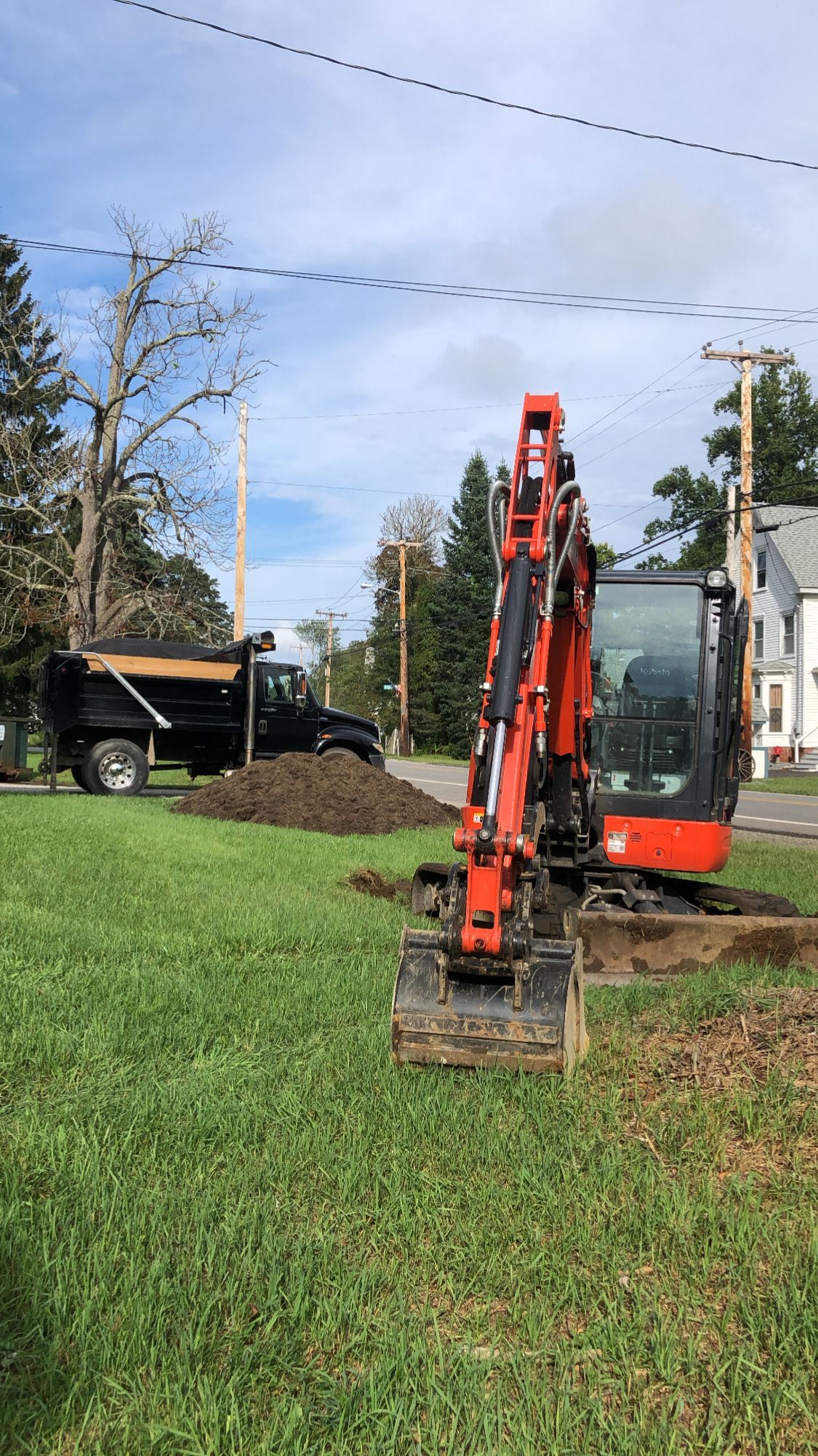 An orange compact excavator sits on a green lawn near a pile of dirt and a black dump truck in a residential area.