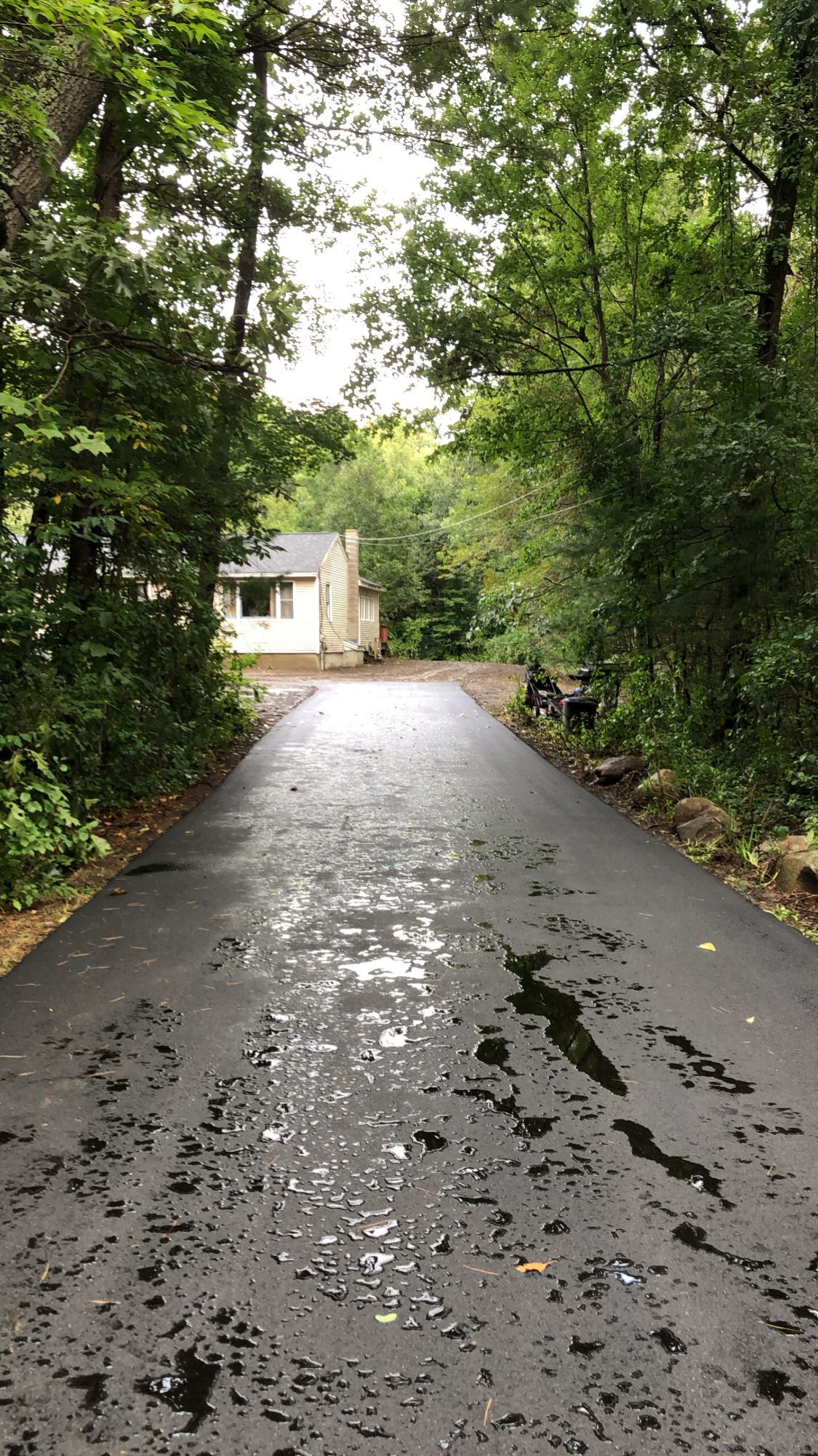 A freshly paved, dark asphalt driveway leads through a wooded area toward a light-colored house in the distance.
