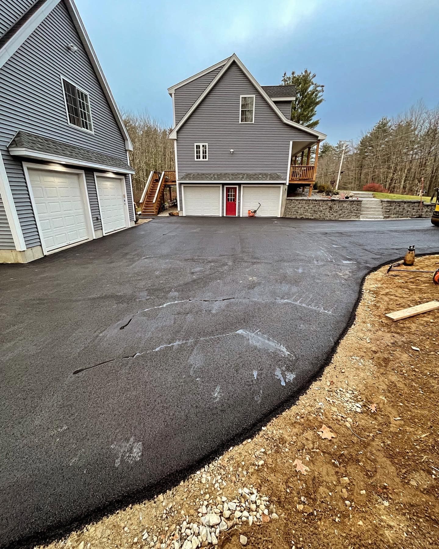 A wide asphalt driveway leads toward a gray two-story house with two garage doors and a red entry door.